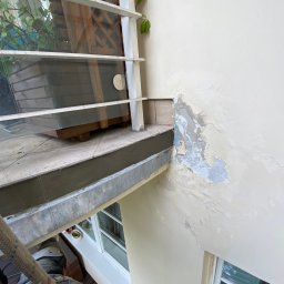 Yury Druhau - Pracownik na linie zabezpieczającej, widoczny z góry, przygotowuje ścianę zewnętrzną budynku do malowania; łuszcząca się farba na elewacji, okno z białą ramą w tle.