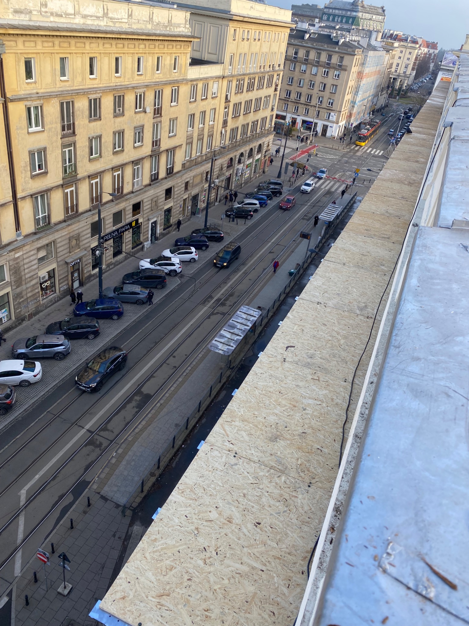 Widok z góry na miejską ulicę z budynkami, tramwajem i samochodami, z fragmentem zabezpieczonej płyty OSB na pierwszym planie, prawdopodobnie z dachu budynku.
