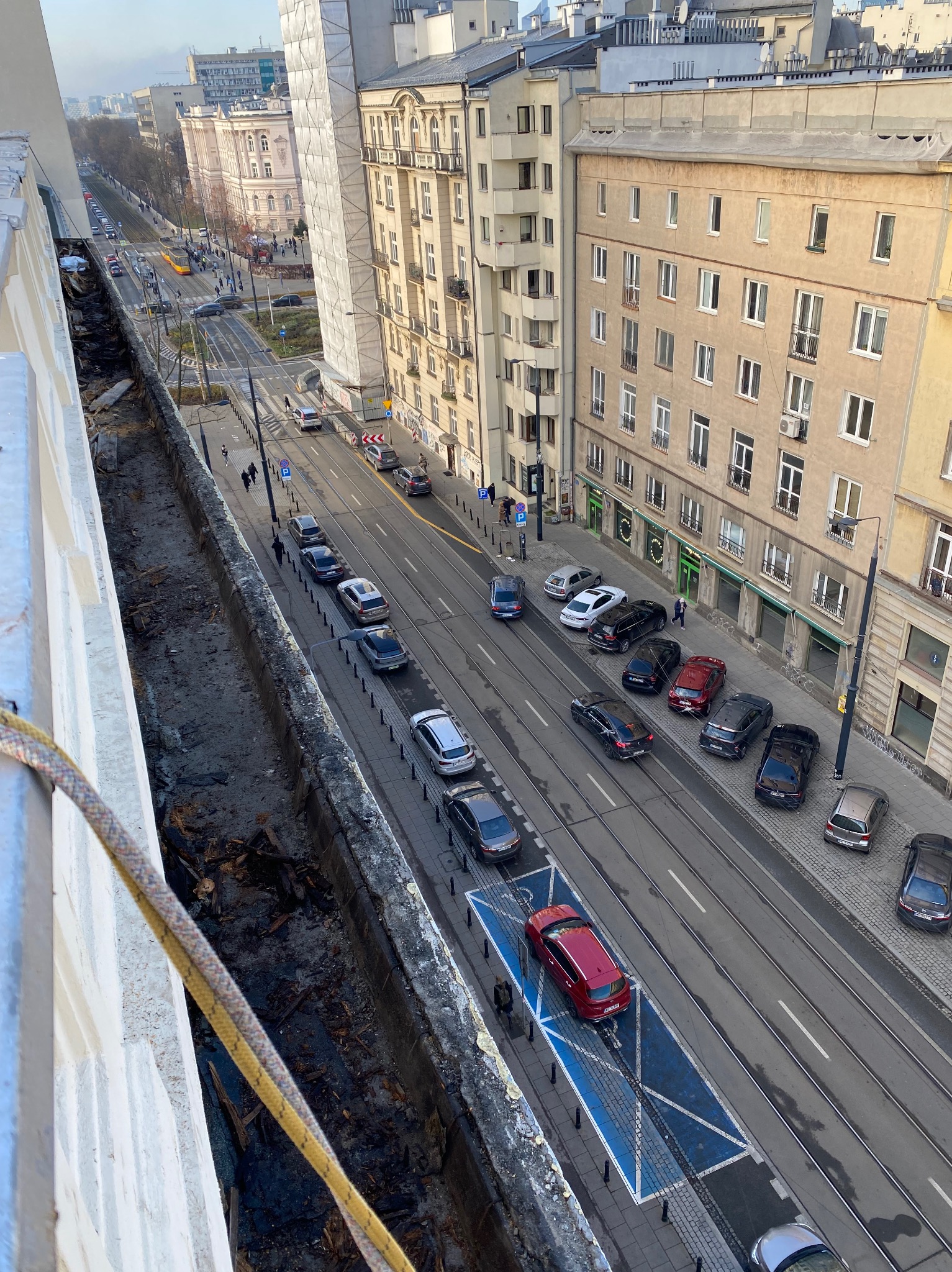 Widok z góry na miejską ulicę z tramwajami i zaparkowanymi samochodami, z fragmentem balkonu po lewej stronie, gdzie widoczne są pozostałości po remoncie i lina zabezpieczająca.