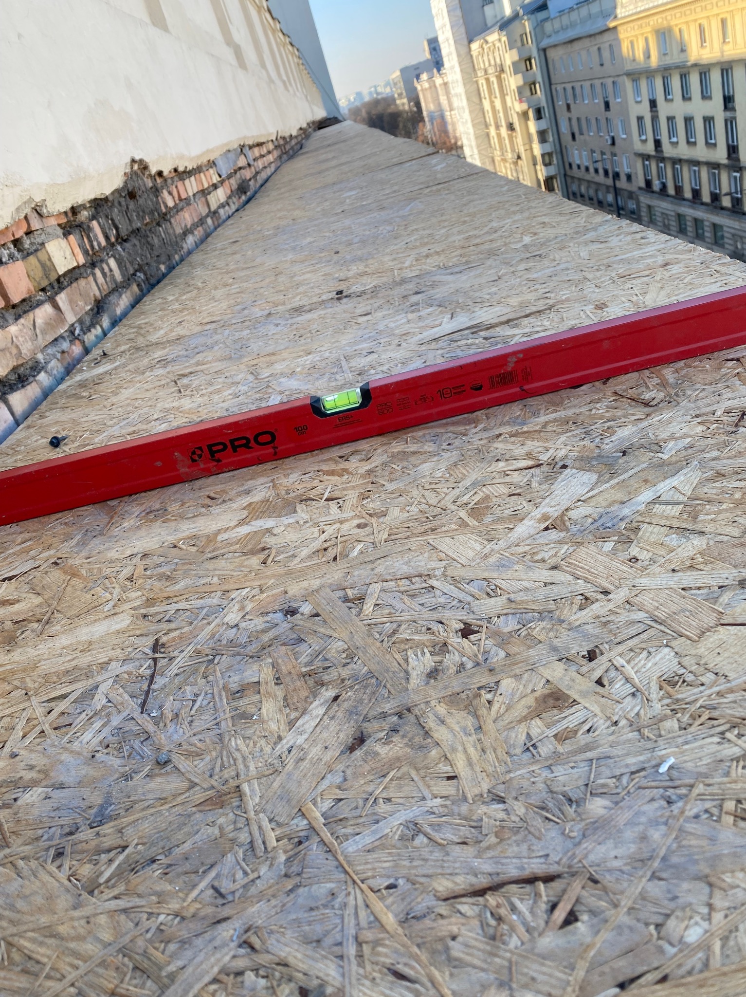 Czerwona poziomica na płycie OSB na dachu budynku w trakcie prac budowlanych, w tle zabudowa miejska.