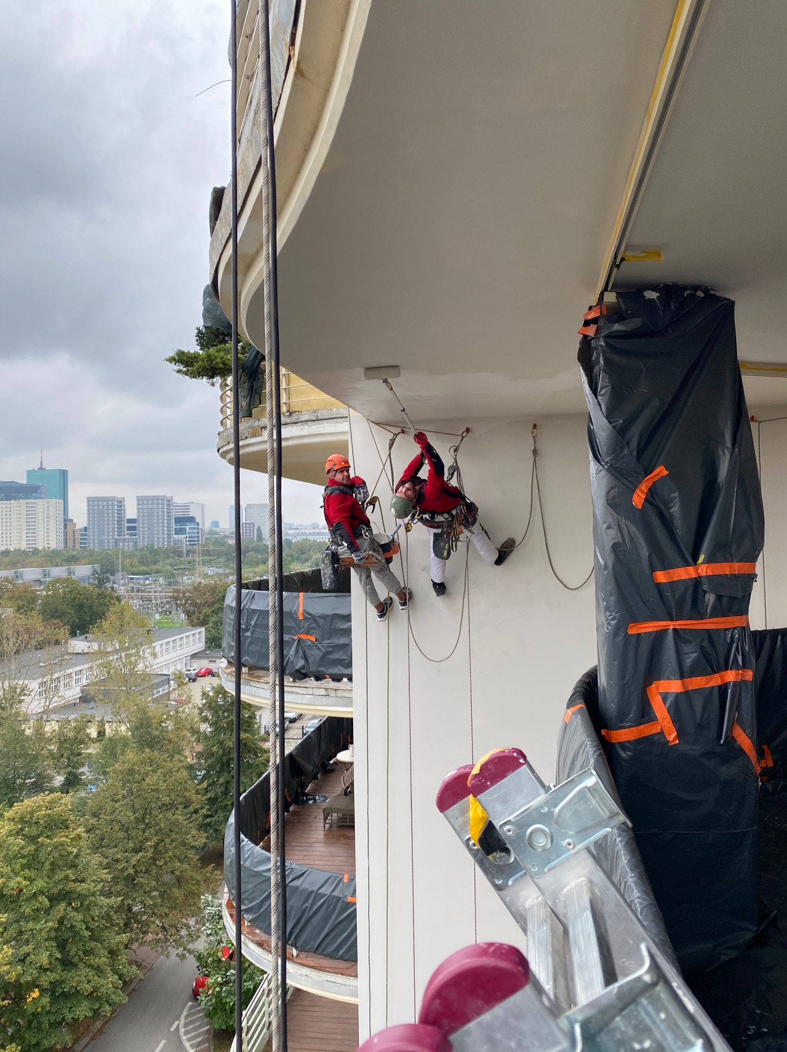 Dwóch pracowników na linach zabezpieczających, w kaskach i kombinezonach, wykonuje prace na wysokości na elewacji budynku; widoczna drabina i osłonięte elementy budynku folią; w tle panorama miasta.