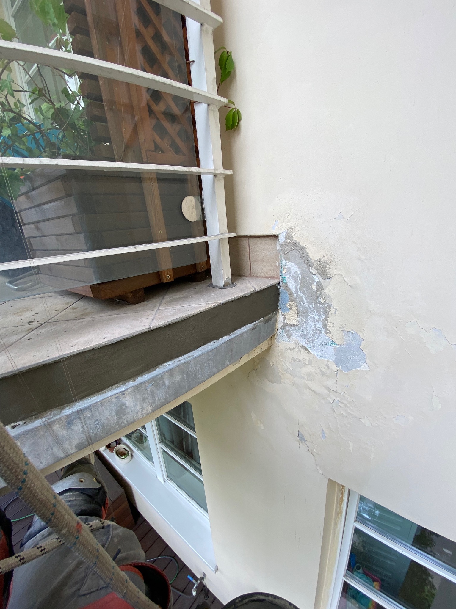 Pracownik na linie zabezpieczającej, widoczny z góry, przygotowuje ścianę zewnętrzną budynku do malowania; łuszcząca się farba na elewacji, okno z białą ramą w tle.