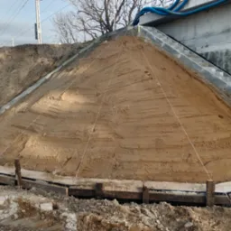 Przygotowanie podłoża: ziemny nasyp uformowany w stożek, otoczony drewnianą konstrukcją, z widocznymi liniami wyznaczającymi kształt. Schody betonowe w tle.