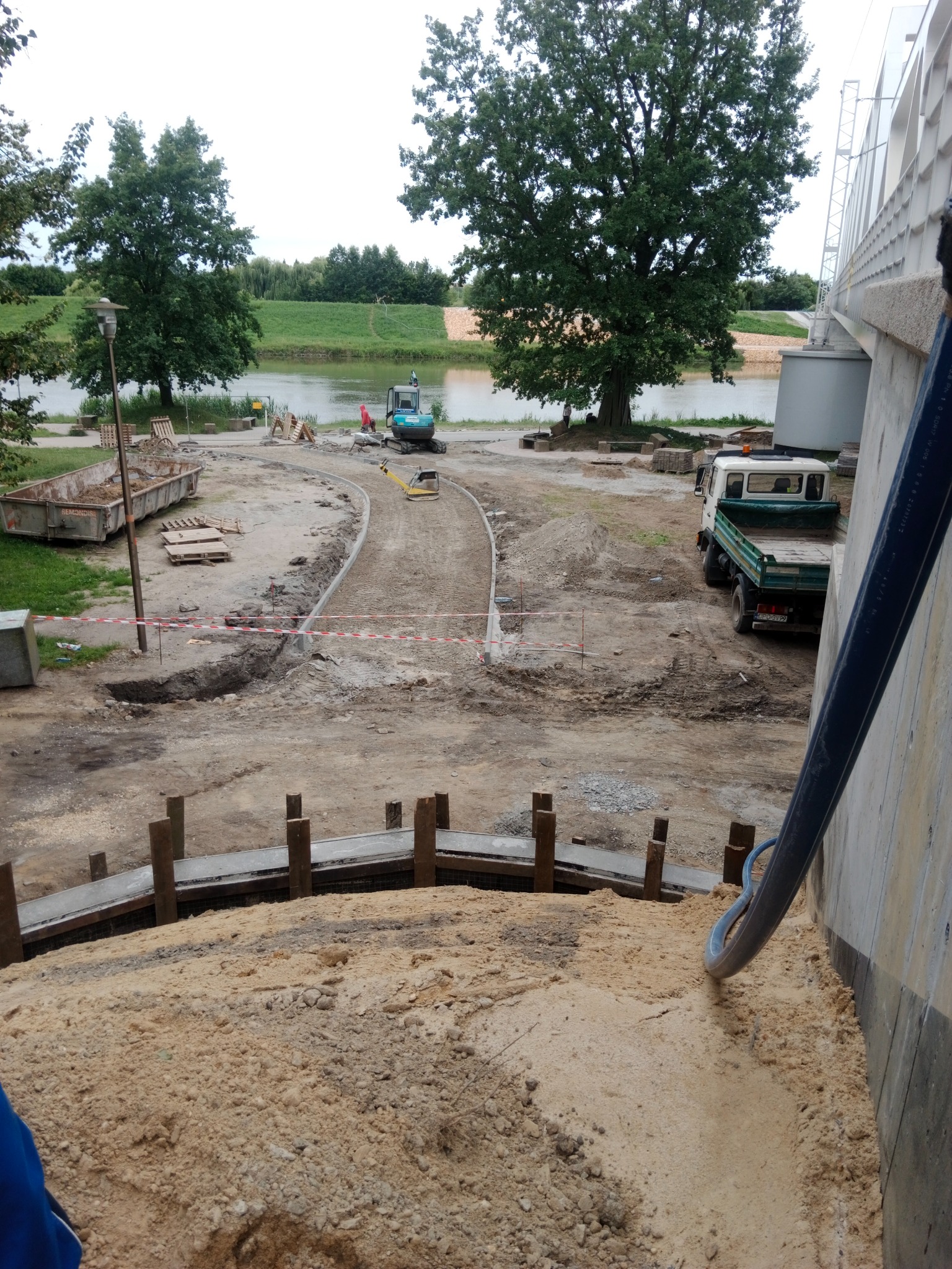 Budowa ścieżki parkowej z wykopem, rurą doprowadzającą piasek, mini koparką w tle. Widać elementy konstrukcyjne i przygotowane podłoże.