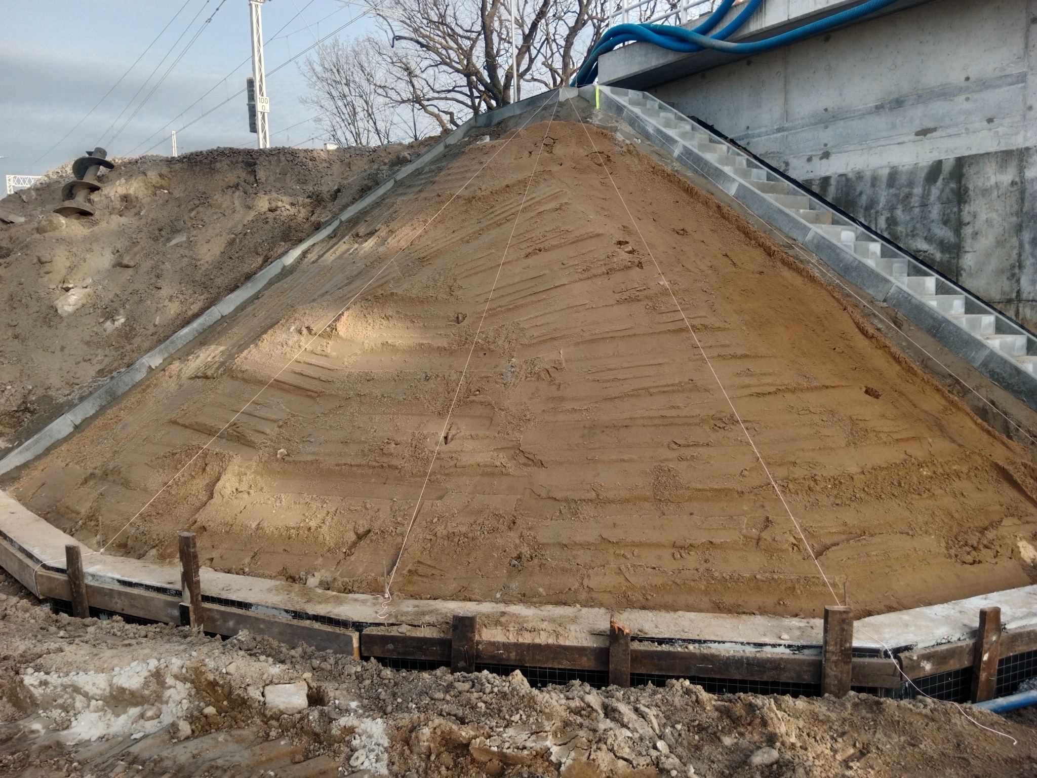 Przygotowanie podłoża: ziemny nasyp uformowany w stożek, otoczony drewnianą konstrukcją, z widocznymi liniami wyznaczającymi kształt. Schody betonowe w tle.