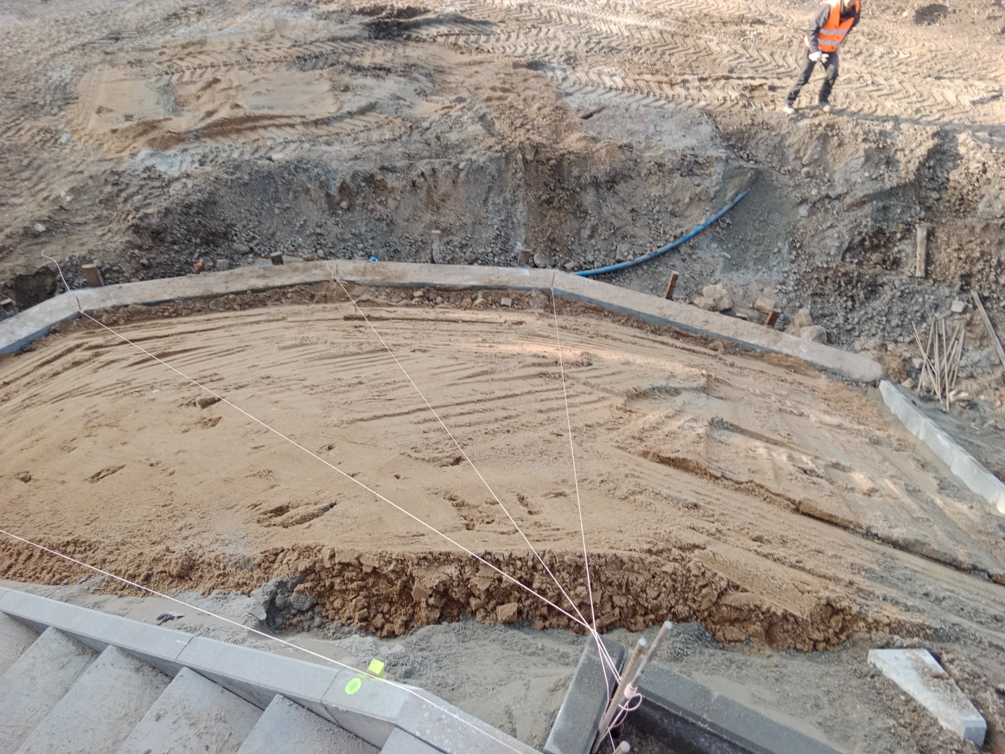 Przygotowanie terenu pod budowę schodów zewnętrznych z kostki brukowej. Widoczne wykopaliska, sznurki wyznaczające poziom i pracownik w oddali.