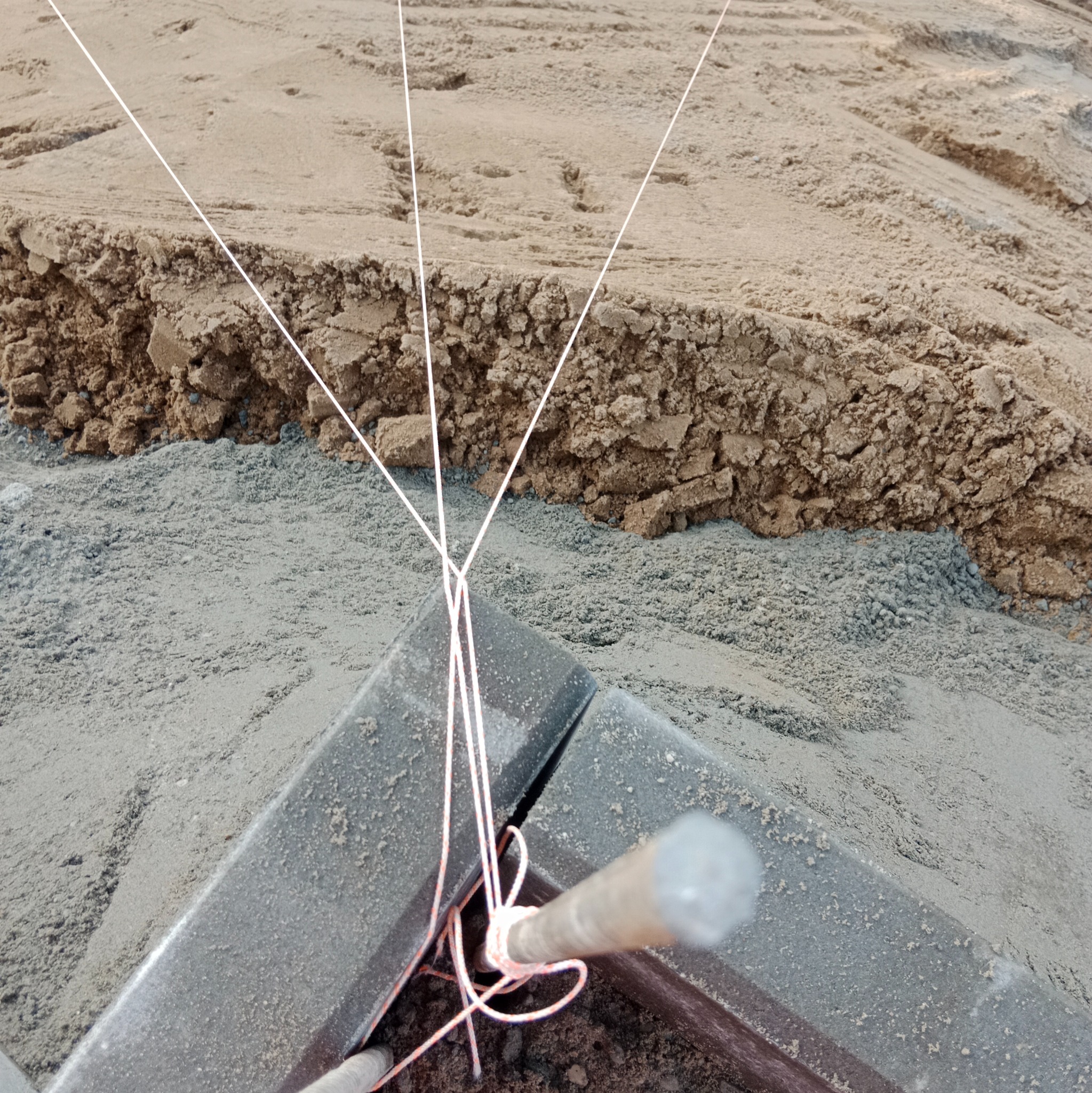 Ustawianie kostki brukowej: sznurki wyznaczają linię, wbity pręt stabilizuje. Podłoże z piasku i ziemi przygotowane pod nawierzchnię.