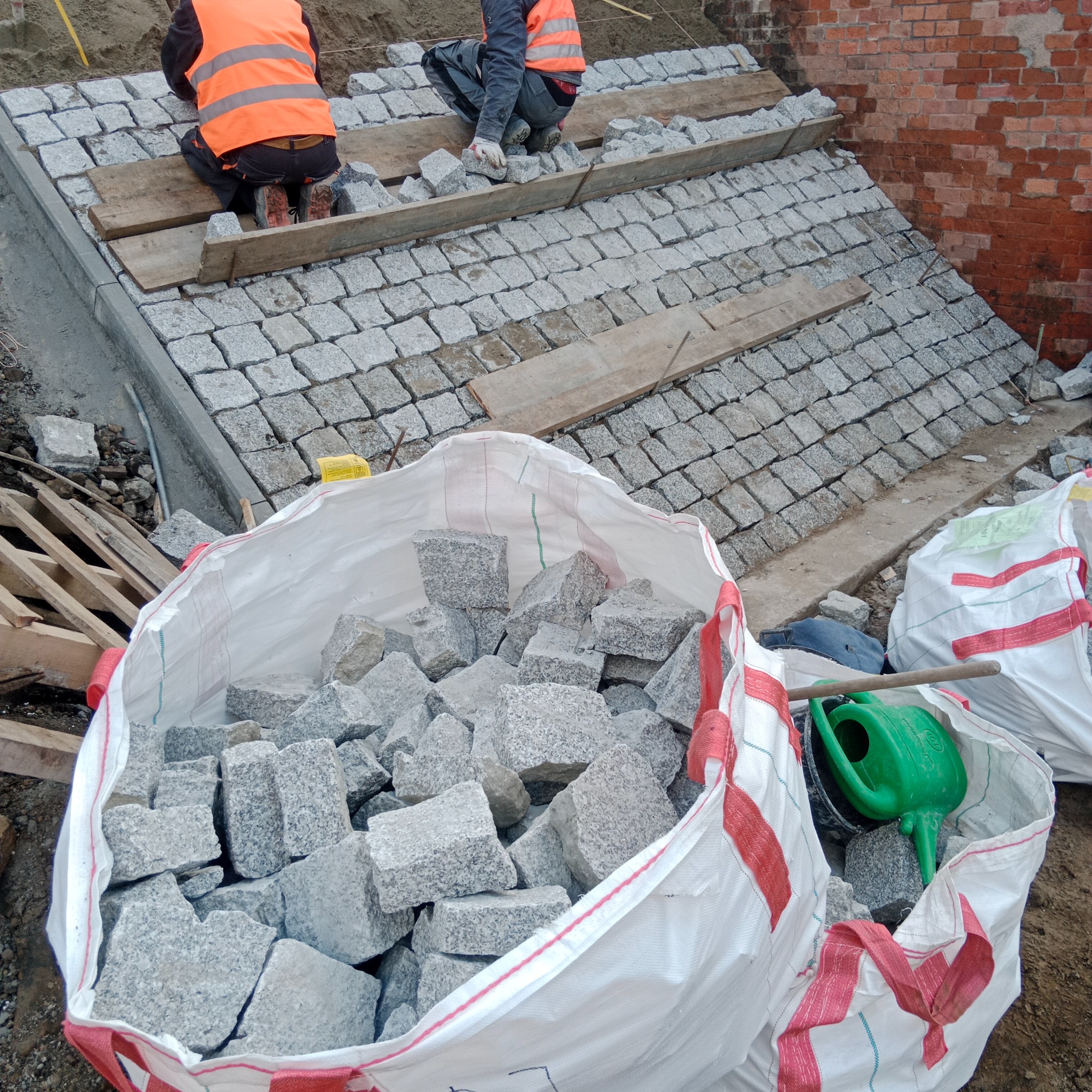 Układanie kostki granitowej na podjeździe. Widoczne worki z kostką, dwóch pracowników w kamizelkach odblaskowych oraz fragment muru z czerwonej cegły w tle.