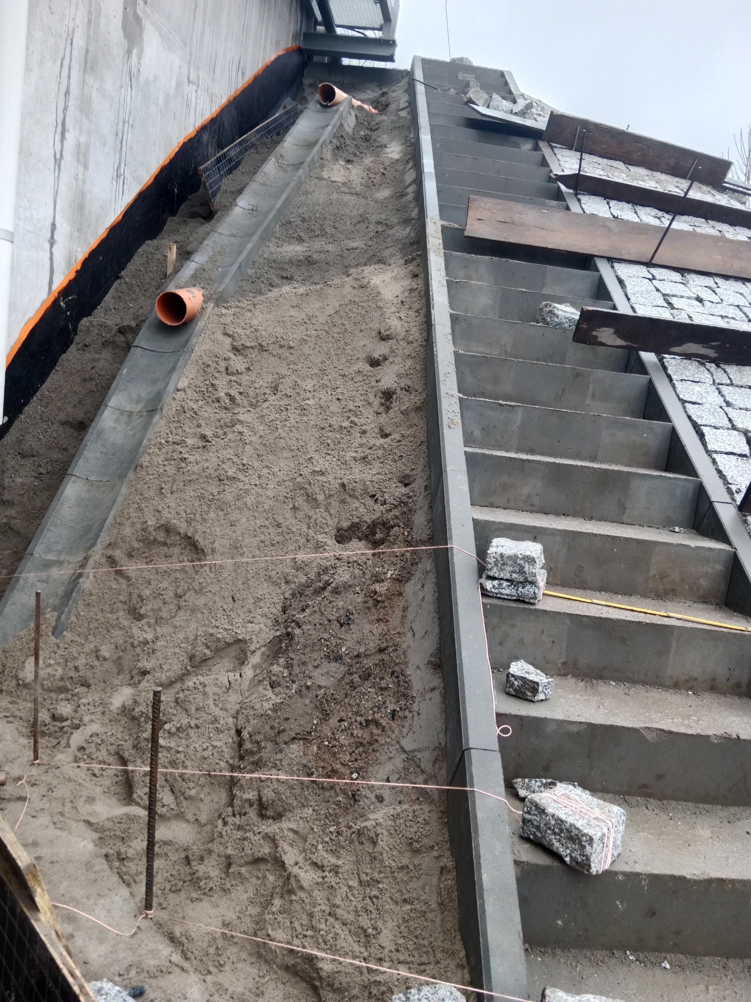 Budowa schodów zewnętrznych z kostki granitowej na podsypce piaskowej. Widoczne rury drenażowe i elementy konstrukcyjne. Kąt ujęcia z dołu.