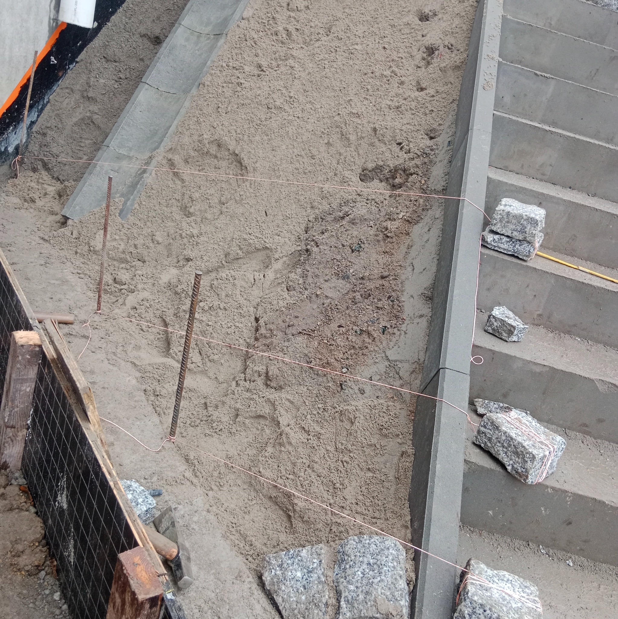Przygotowanie podłoża pod schody z kostki granitowej. Widoczne sznurki wyznaczające poziom, piasek, obrzeża i kostka granitowa. Narzędzia pracy w tle.