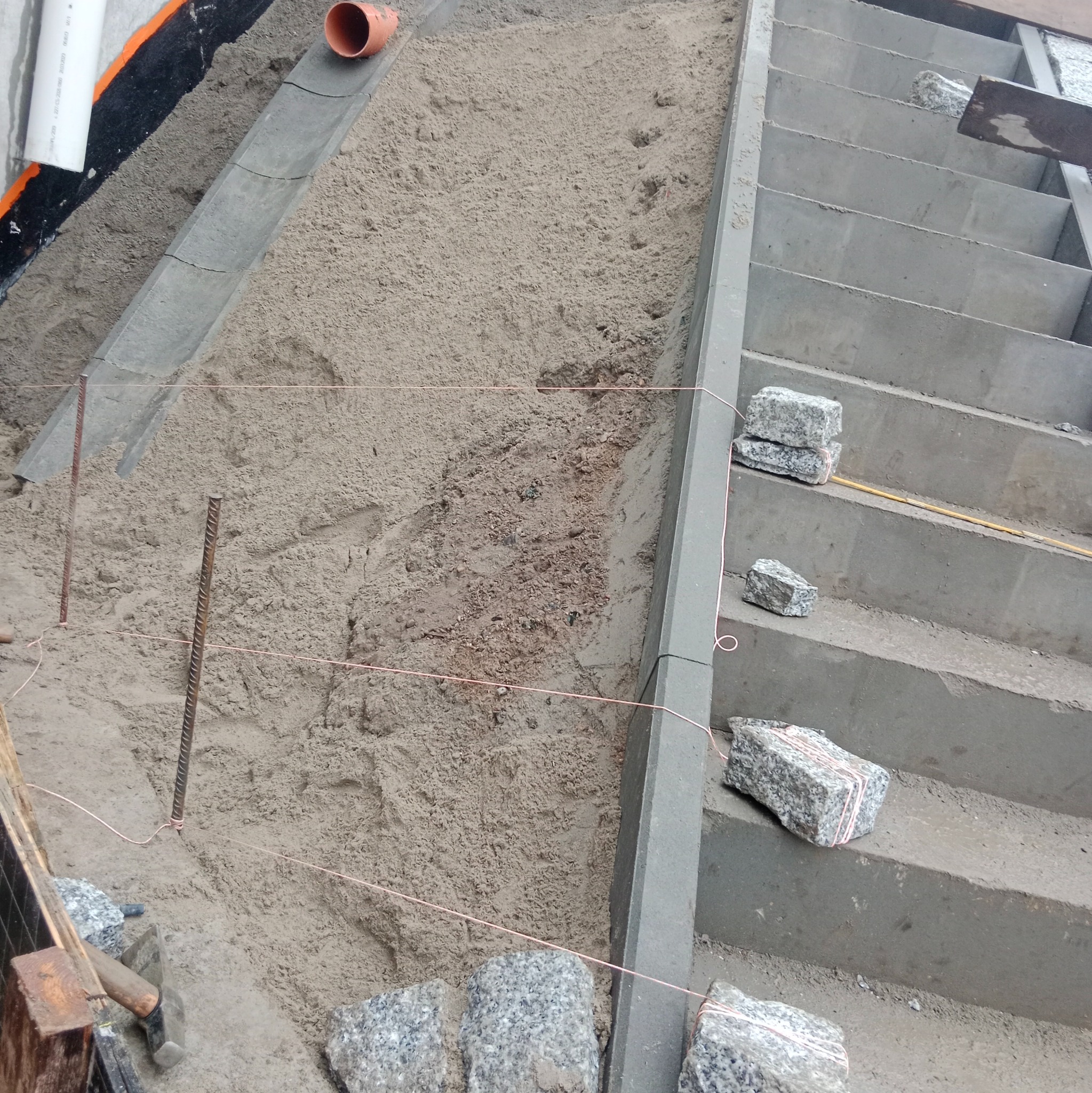Przygotowanie podłoża pod schody z kostki granitowej. Widoczne sznurki wyznaczające poziom, podsypka piaskowa i częściowo ułożone kamienie.