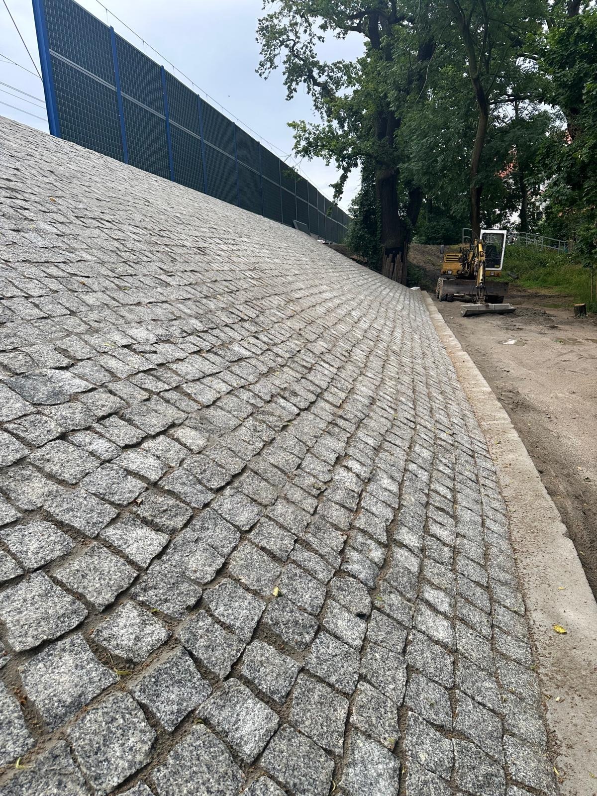 Skarpa wyłożona kostką granitową w szarym odcieniu, z widocznym małym traktorem w tle i ekranem akustycznym. Układ ukośny kostki, widok z dołu.