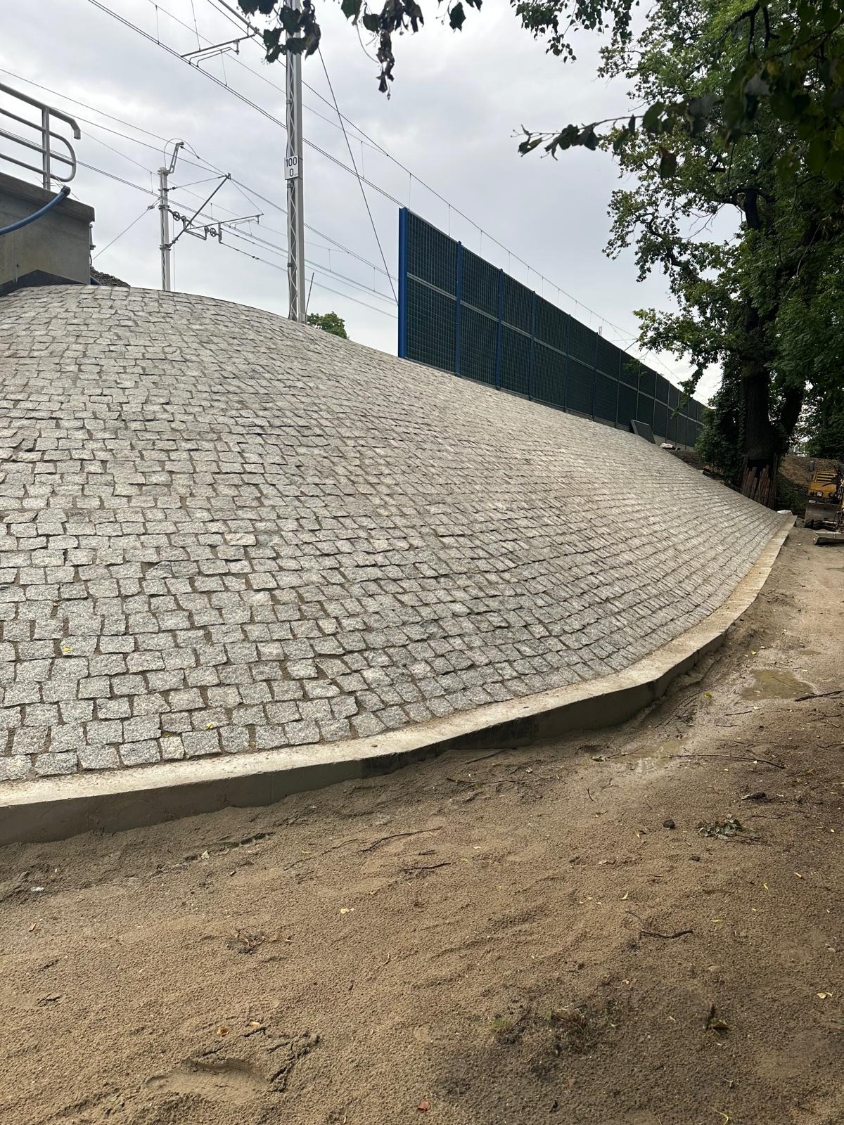 Granitowa skarpa przy torach kolejowych, zabezpieczona betonową obwódką i ekranem akustycznym. Widoczne drzewa i słupy trakcyjne.