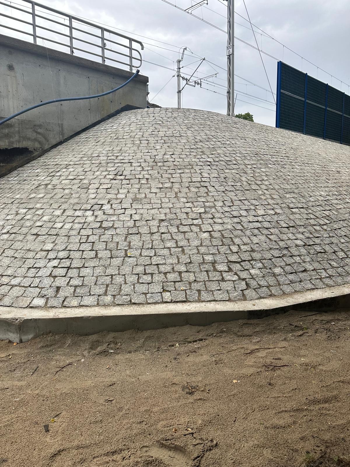 Kopuła z kostki granitowej pod wiaduktem kolejowym, widoczna z poziomu gruntu. Kostka ułożona w regularny wzór, tło stanowią elementy infrastruktury kolejowej.