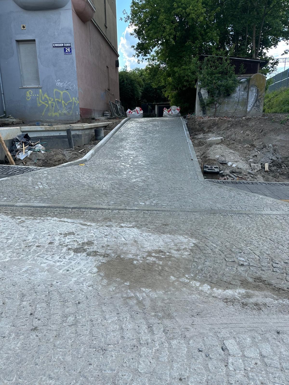 Świeżo ułożona, mokra kostka granitowa tworząca podjazd. Widoczne worki z materiałem i budynek z graffiti w tle. Praca w toku.