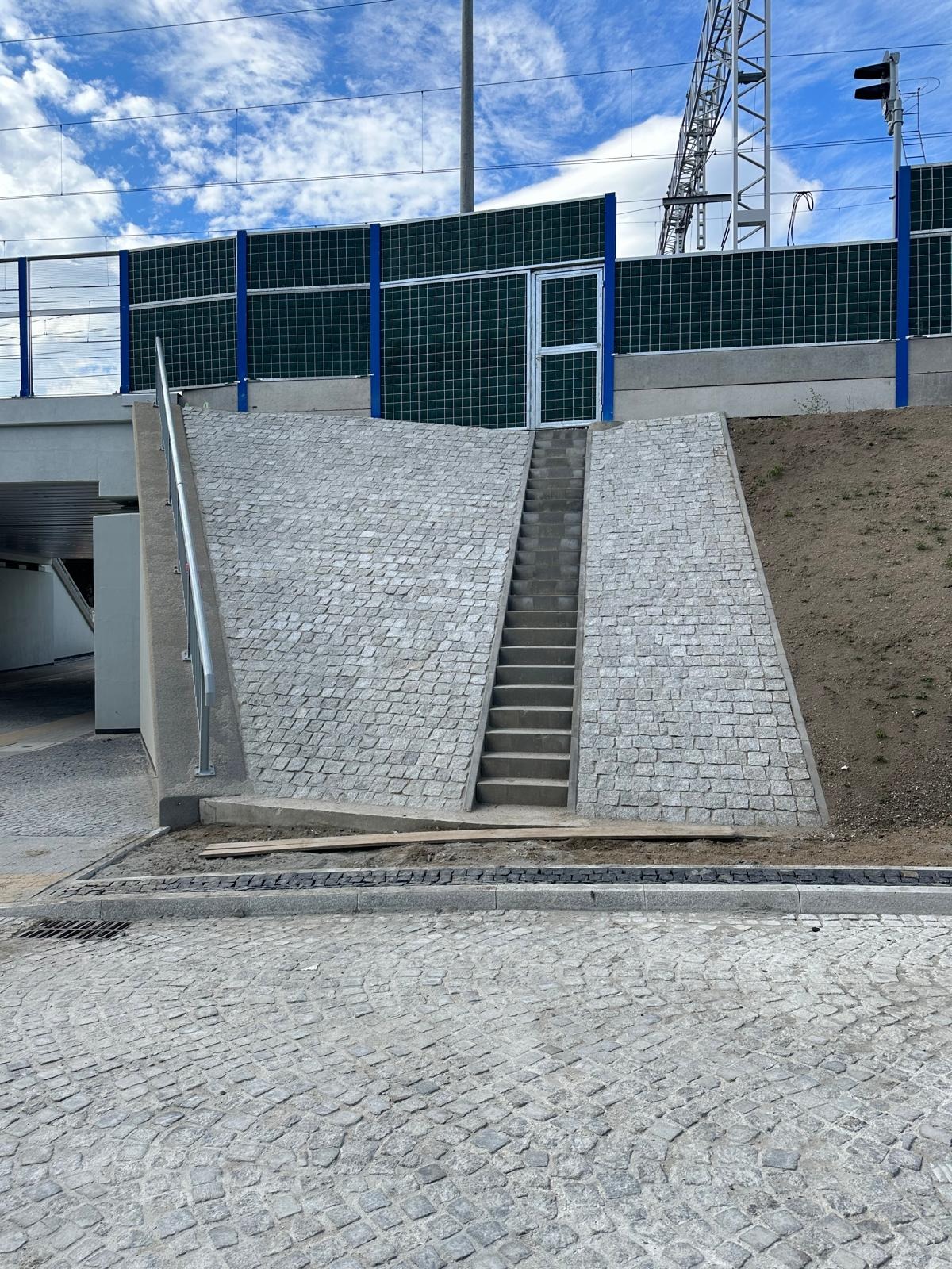 Schody z kostki granitowej prowadzące do wiaduktu, otoczone murem z tego samego materiału. Widoczny fragment chodnika z kostki i metalowa barierka.