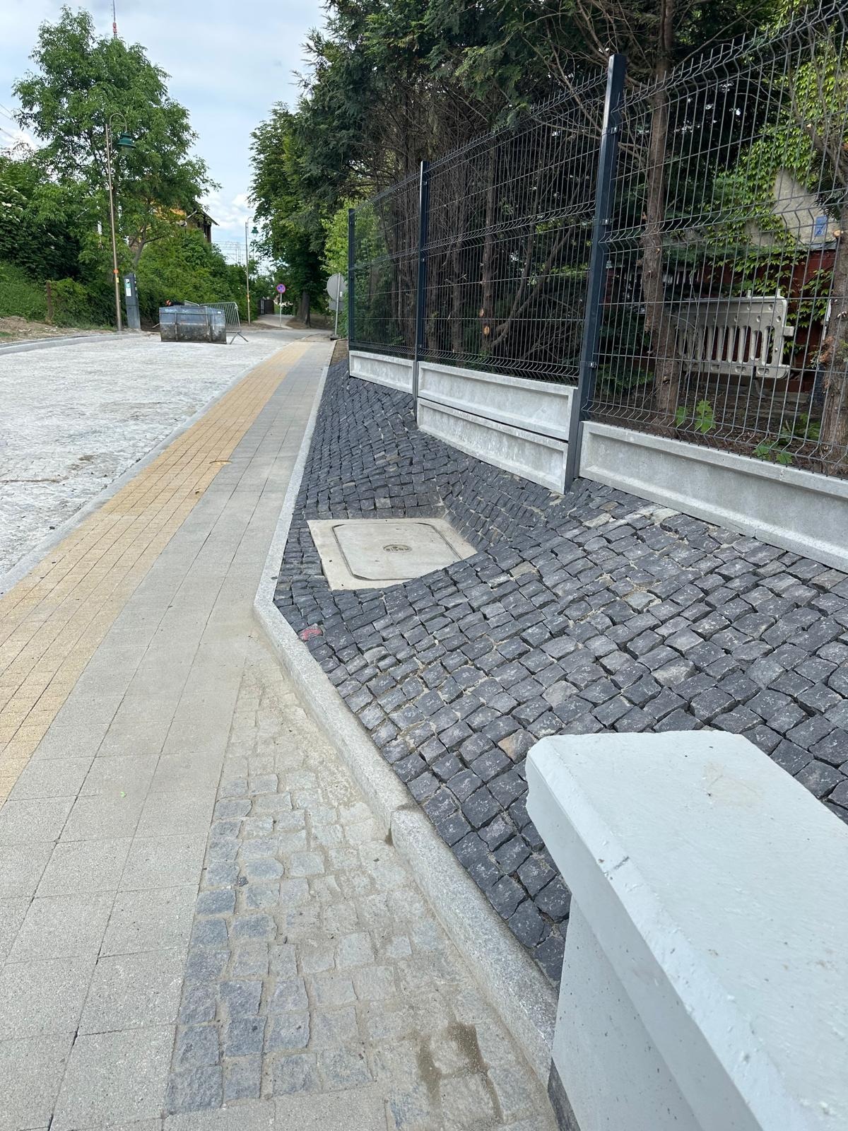 Ukończony fragment chodnika z kostki brukowej w odcieniach szarości i żółci, oraz skarpa z kostki granitowej zabezpieczona siatką ogrodzeniową.