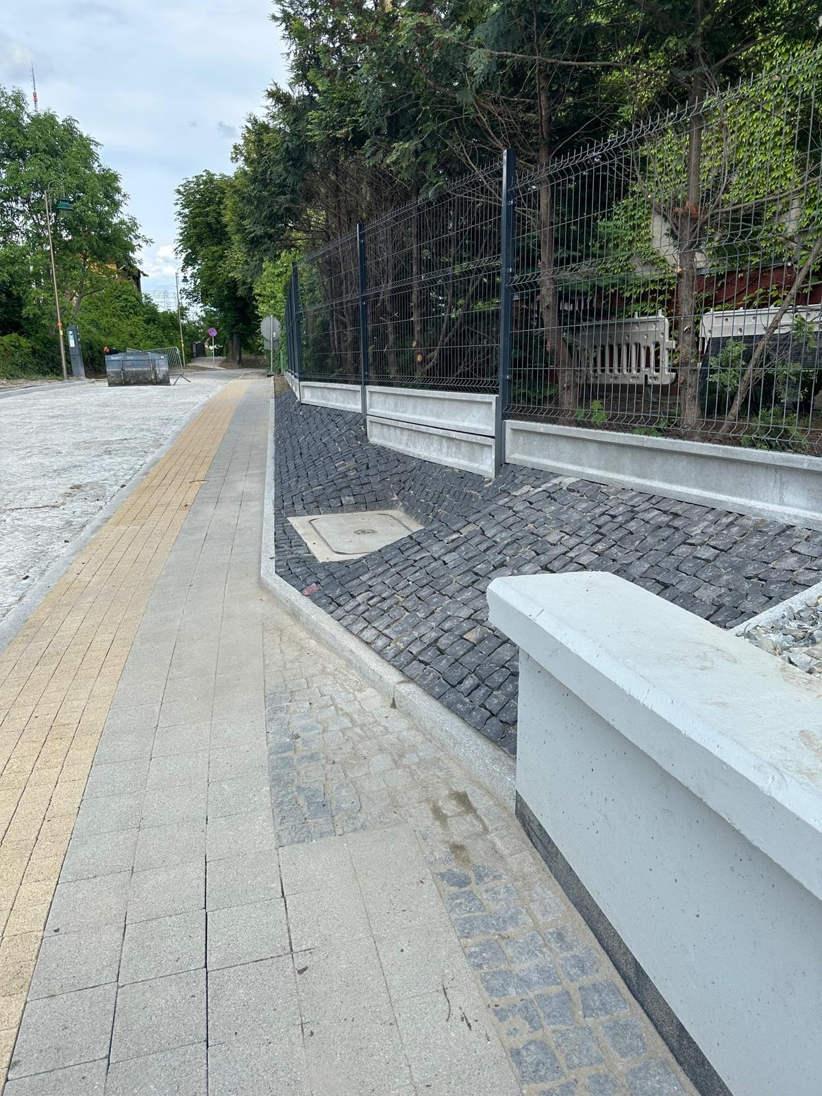 Ukończona nawierzchnia z kostki granitowej wzdłuż chodnika, zabezpieczona betonową ścianką oporową i ogrodzeniem panelowym na skarpie. Widok na ulicę.