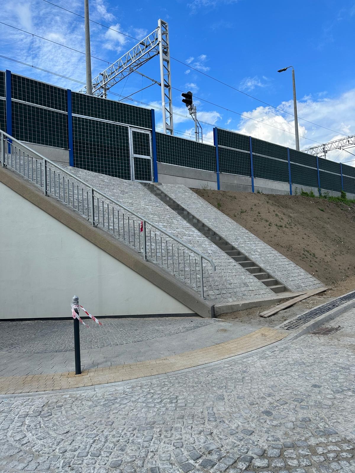 Granitowe schody z metalową barierką prowadzą pod wiaduktem kolejowym. Tło: ekran akustyczny z siatki i błękitne niebo.