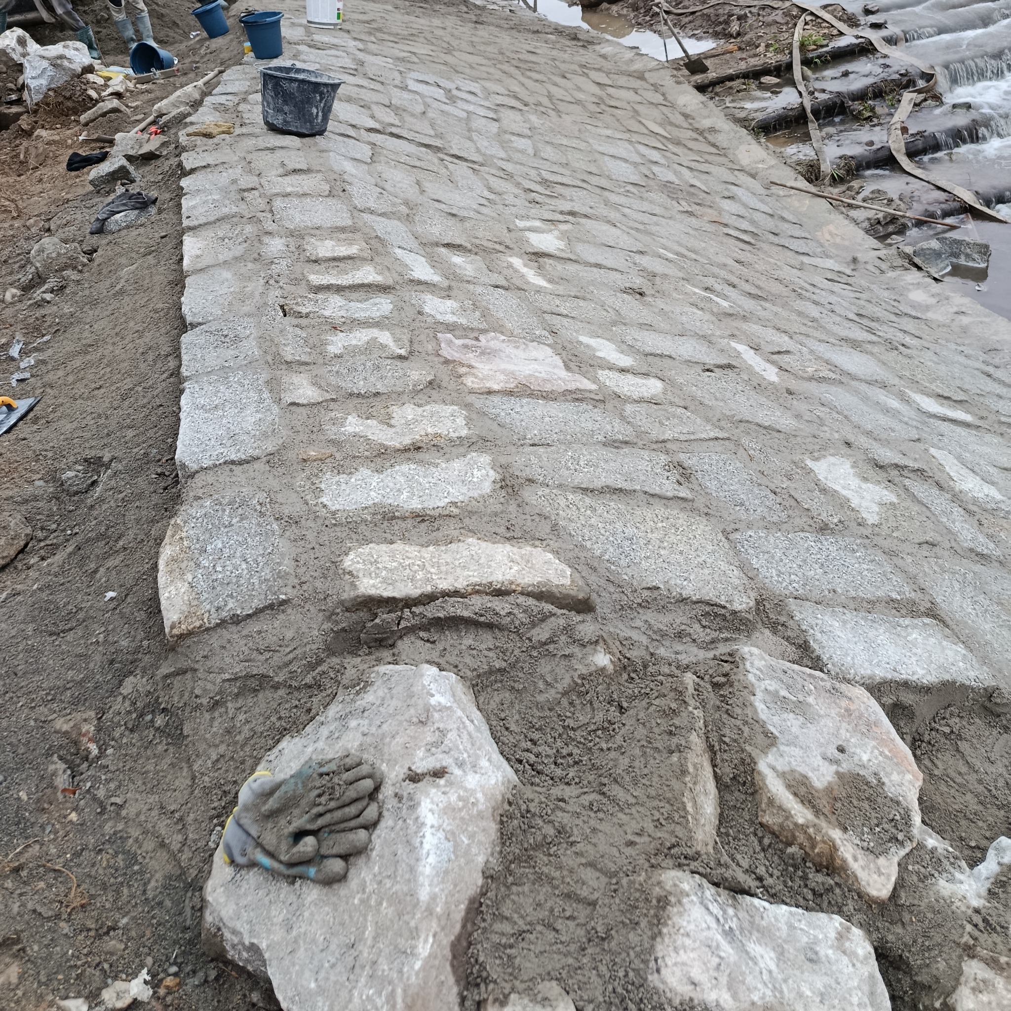 Układanie kostki granitowej na skarpie przy rzece. Widoczne narzędzia, kubeł i rękawice robocze. Kąt ujęcia z dołu podkreśla nachylenie terenu i etap prac.