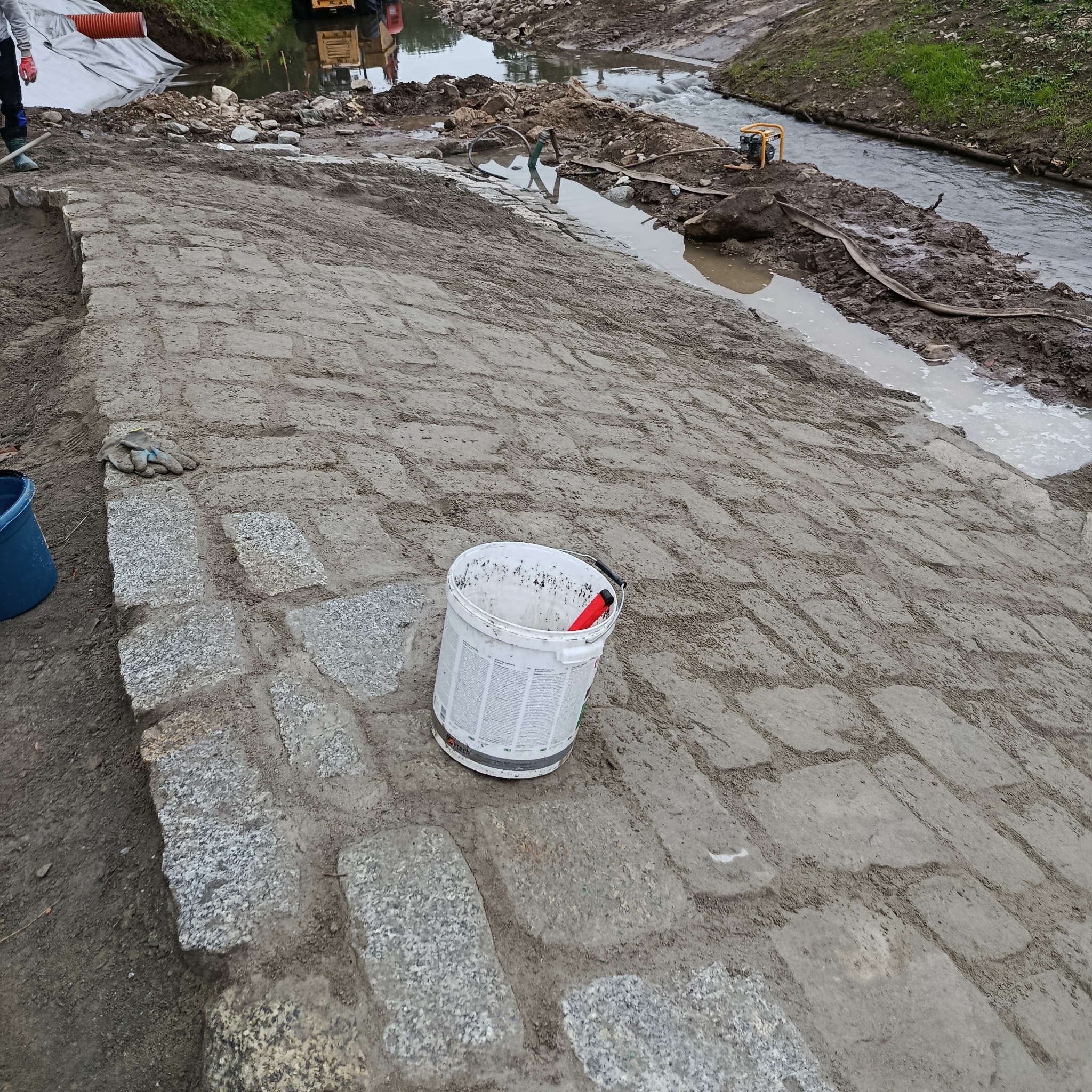 Układanie kostki granitowej w Opolu, wzmocnienie brzegu rzeki. Widoczny biały kubeł, rękawice robocze i częściowo ułożona nawierzchnia z kostki.