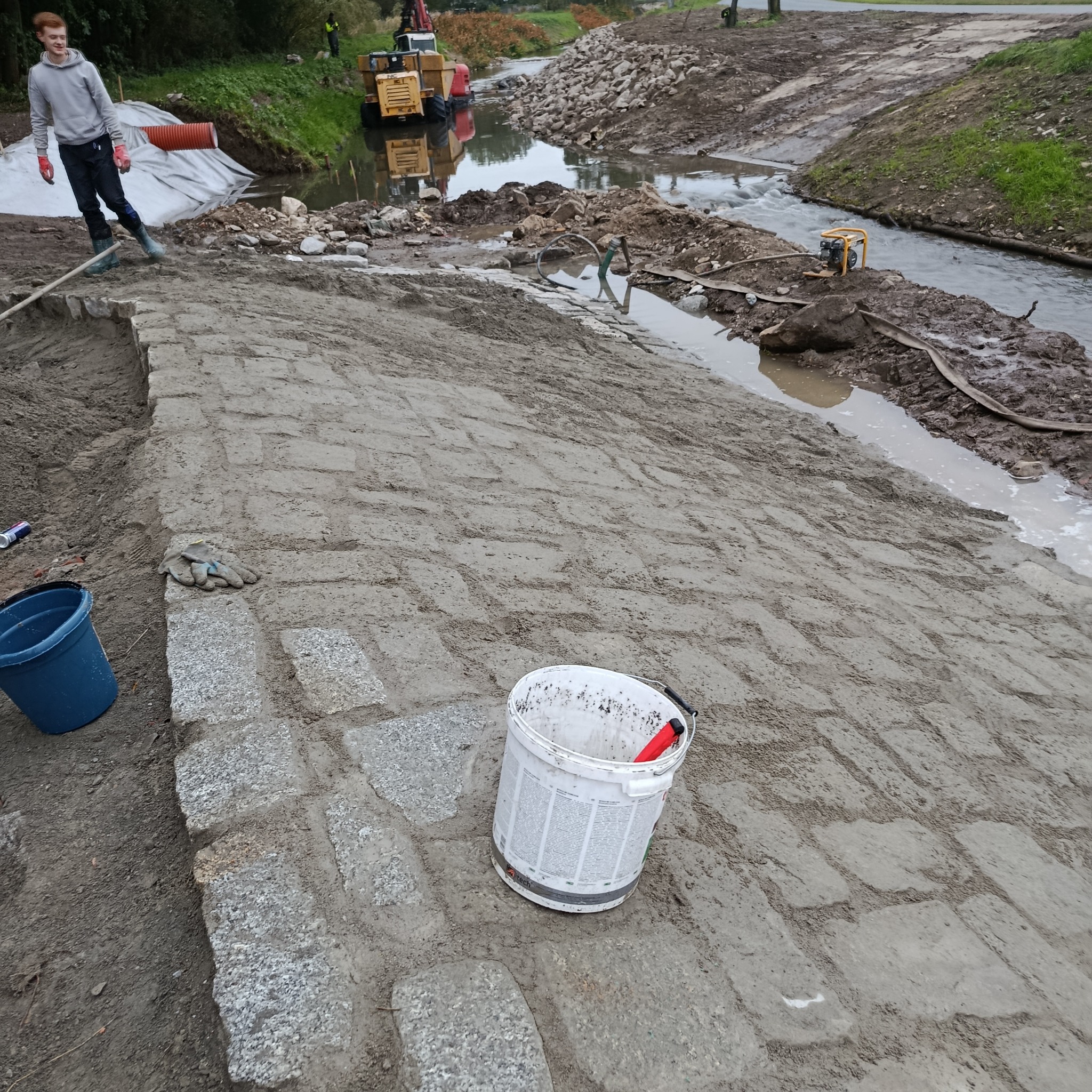 Układanie kostki granitowej na brzegu rzeki, z widocznym sprzętem budowlanym i pracownikiem. Brzeg rzeki wzmacniany kamieniami. W tle drzewa i roślinność.