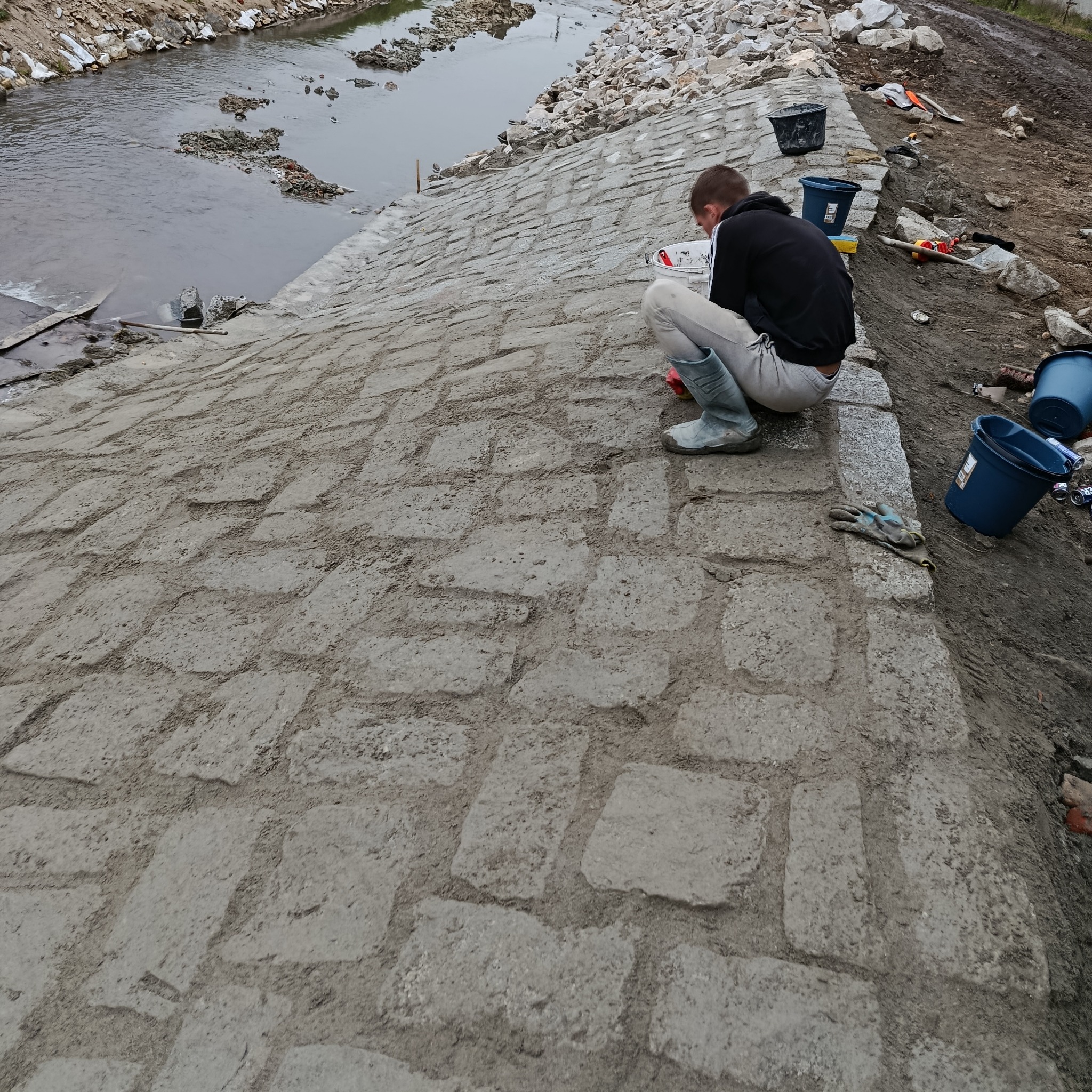 Układanie kamiennej nawierzchni skarpy rzecznej, pracownik w roboczych butach, widoczne narzędzia i rzeka w tle. Prace ziemne i hydrotechniczne w toku.
