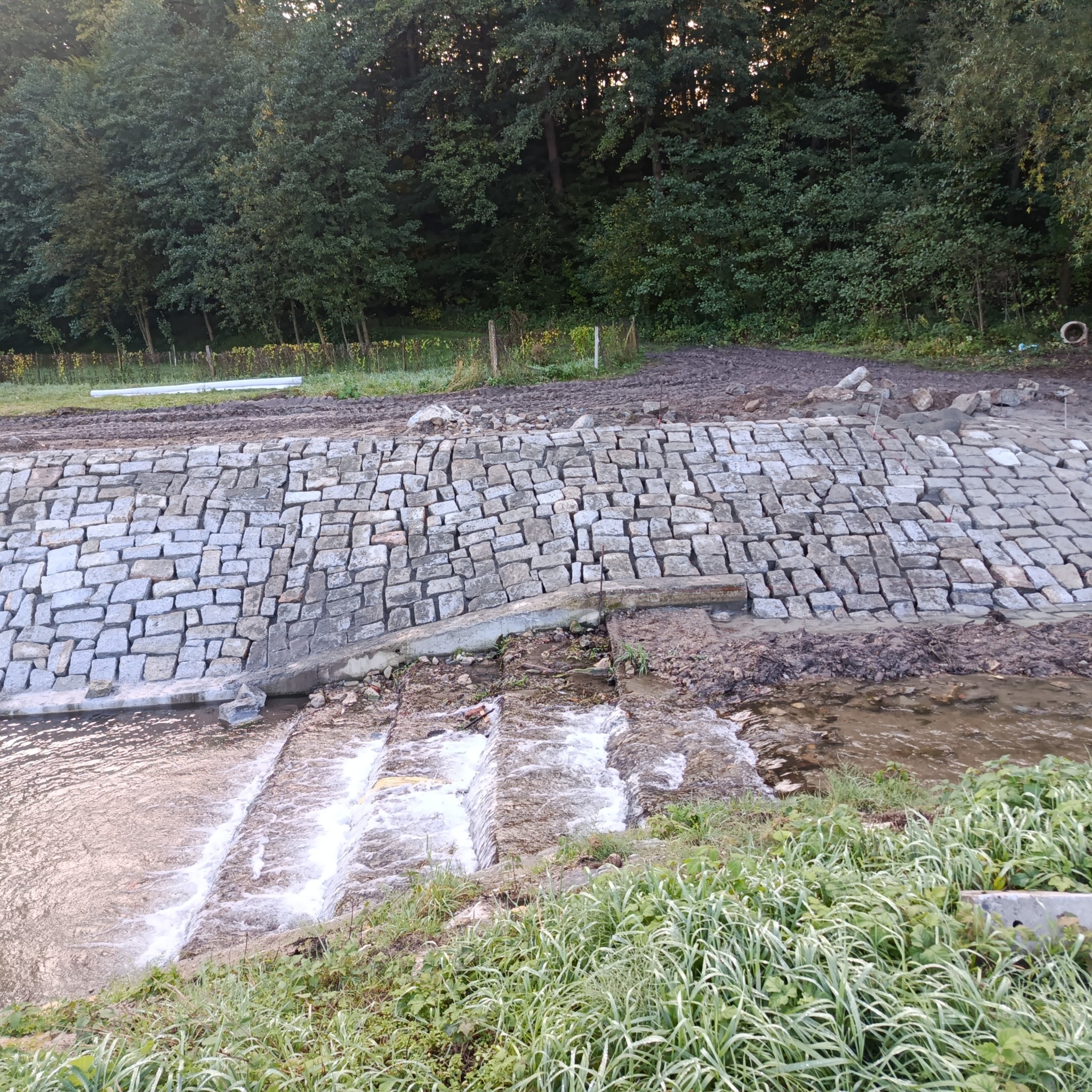 Ułożona kostka granitowa tworząca umocnienie brzegu cieku wodnego. Kamienna konstrukcja z kaskadami na tle zieleni drzew. Woda przepływa przez kamienne stopnie.