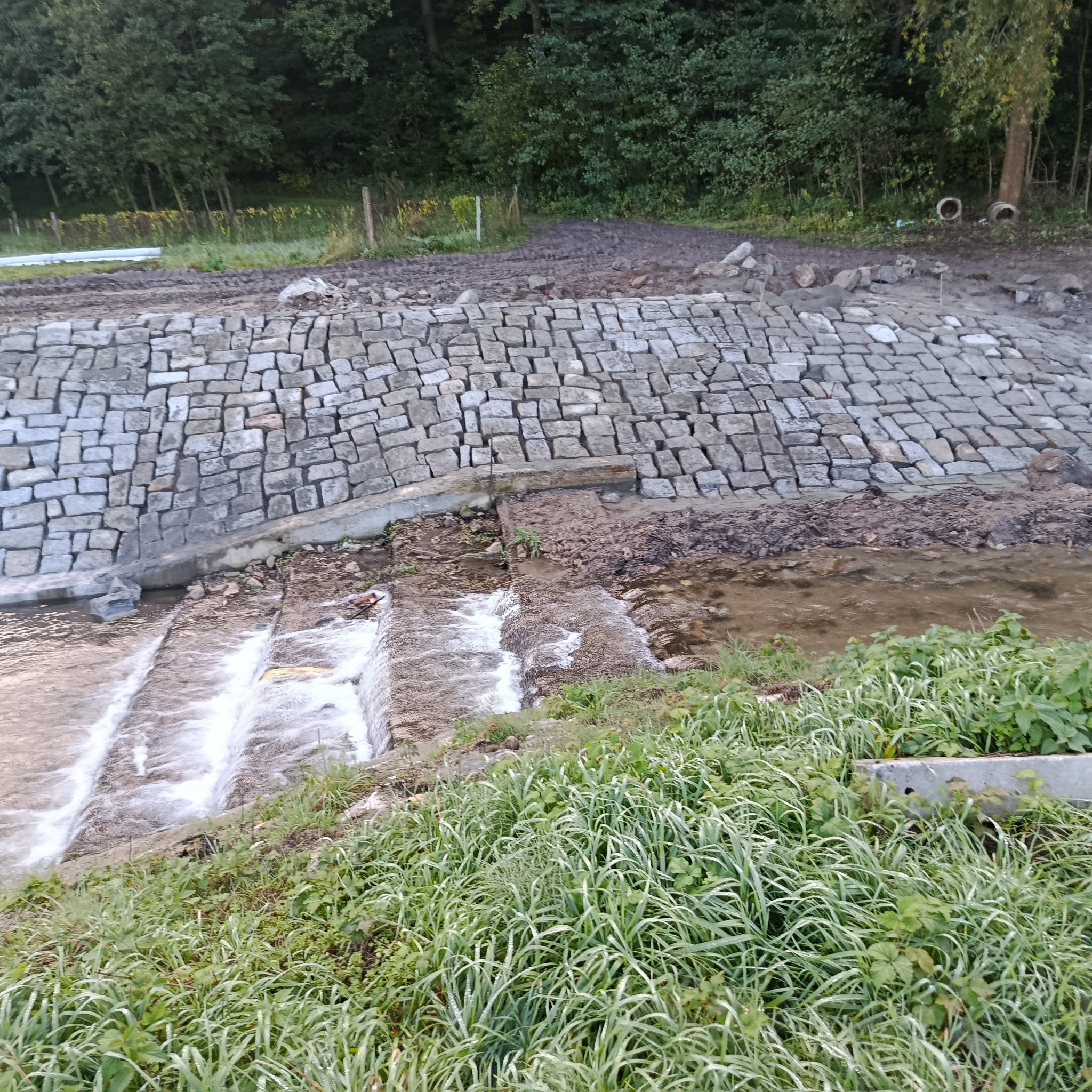Ułożona z szarej kostki granitowej skarpa rzeki, z widocznymi kaskadami wody i zielenią w tle. Prace brukarskie przy regulacji rzeki w naturalnym otoczeniu.