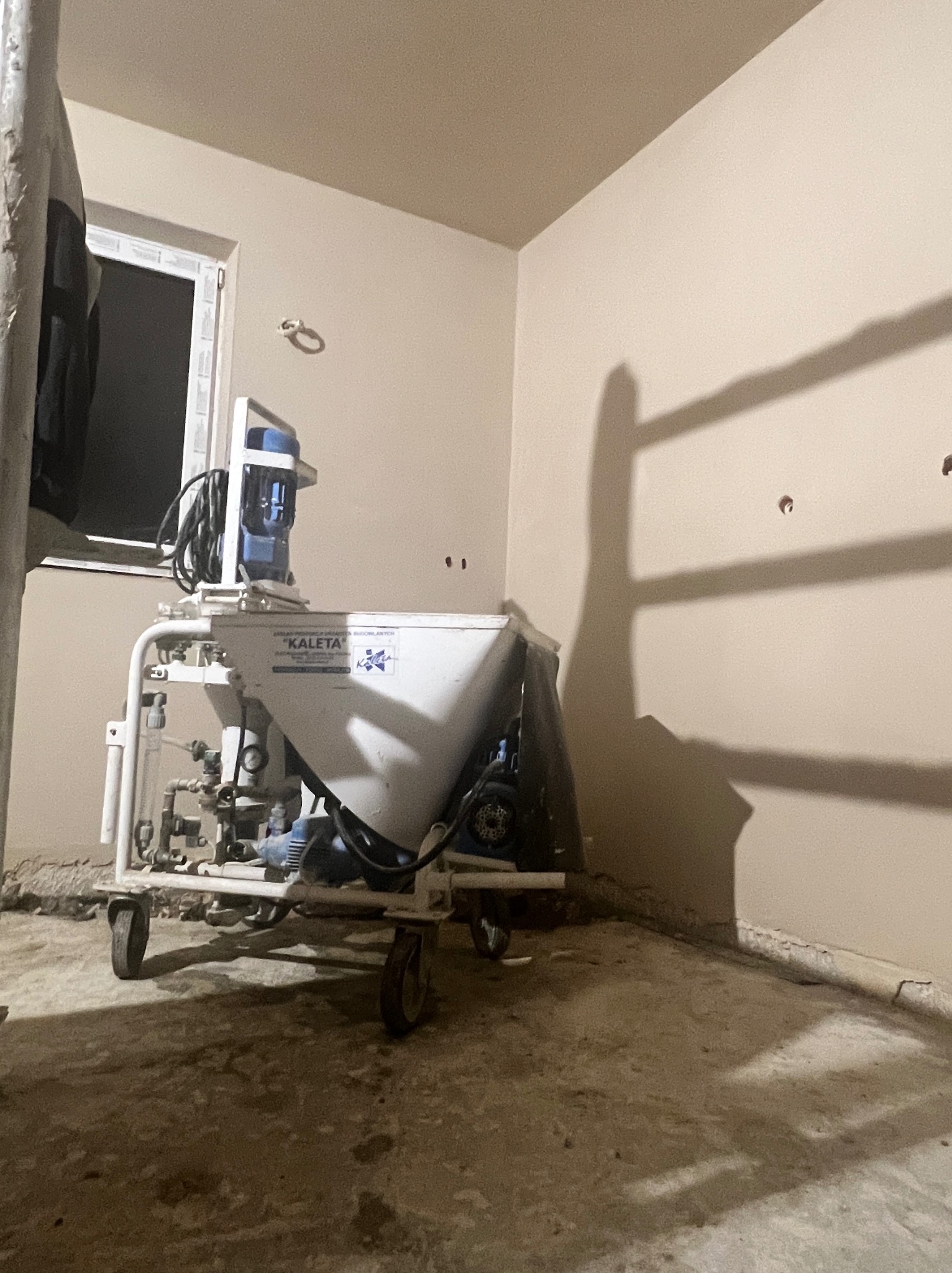 Wnętrze po tynkowaniu maszynowym, widoczna maszyna 'Kaleta' i gładkie, jasne ściany. Ślady po otworach na ścianie. Podłoga surowa, przygotowana do dalszych prac.