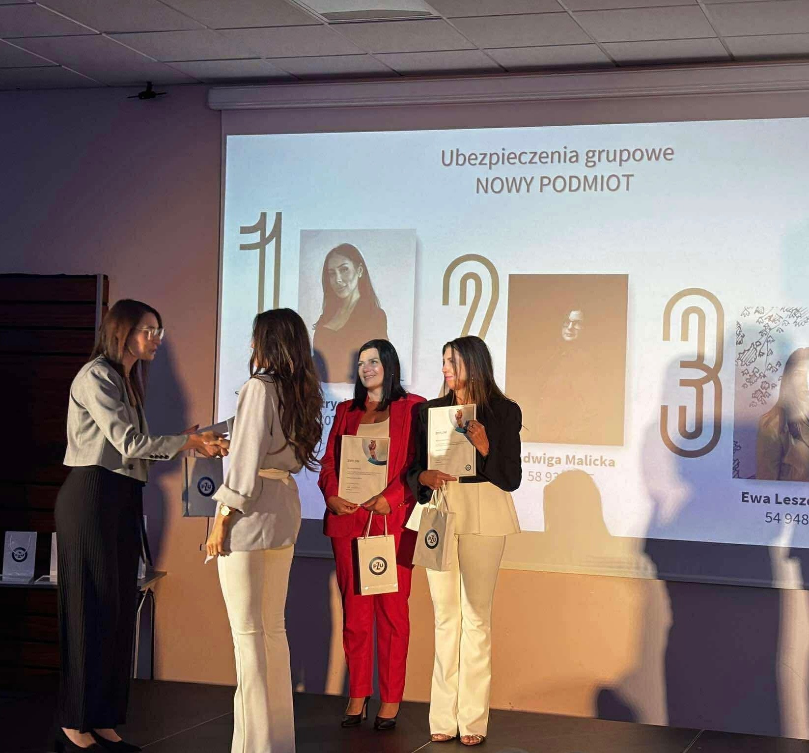 Wręczenie dyplomów i toreb z logo PZU podczas prezentacji 'Ubezpieczenia grupowe – nowy podmiot'. Kobiety w garniturach na tle ekranu z numerami i portretami.