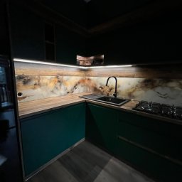 Studio Szkła Rzeszów - Nowoczesna kuchnia z ciemnozielonymi szafkami i blatem imitującym drewno, z podświetleniem LED nad blatem i ozdobną ścianą nad blatem w odcieniach brązu i beżu.