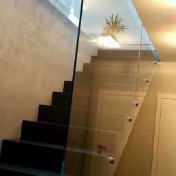 Studio Szkła Rzeszów - Nowoczesne schody z czarnymi stopniami zabezpieczone szklaną balustradą mocowaną punktowo, widok na górny poziom z rośliną doniczkową.