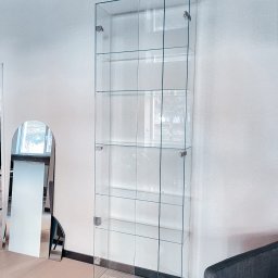 Studio Szkła Rzeszów - Przezroczysta, szklana witryna z półkami i drzwiczkami, zamontowana w nowoczesnym wnętrzu, obok lustra w kształcie łuku.