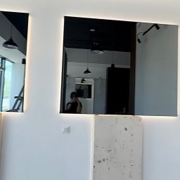 Studio Szkła Rzeszów - Dwa duże, prostokątne lustra z podświetleniem LED na białej ścianie, odbijające wnętrze pomieszczenia z widocznymi lampami sufitowymi i sylwetką osoby robiącej zdjęcie.
