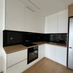 Nowoczesna kuchnia z białymi szafkami, drewnianym blatem i czarnym panelem szklanym na ścianie, widok na zabudowaną płytę indukcyjną i piekarnik.