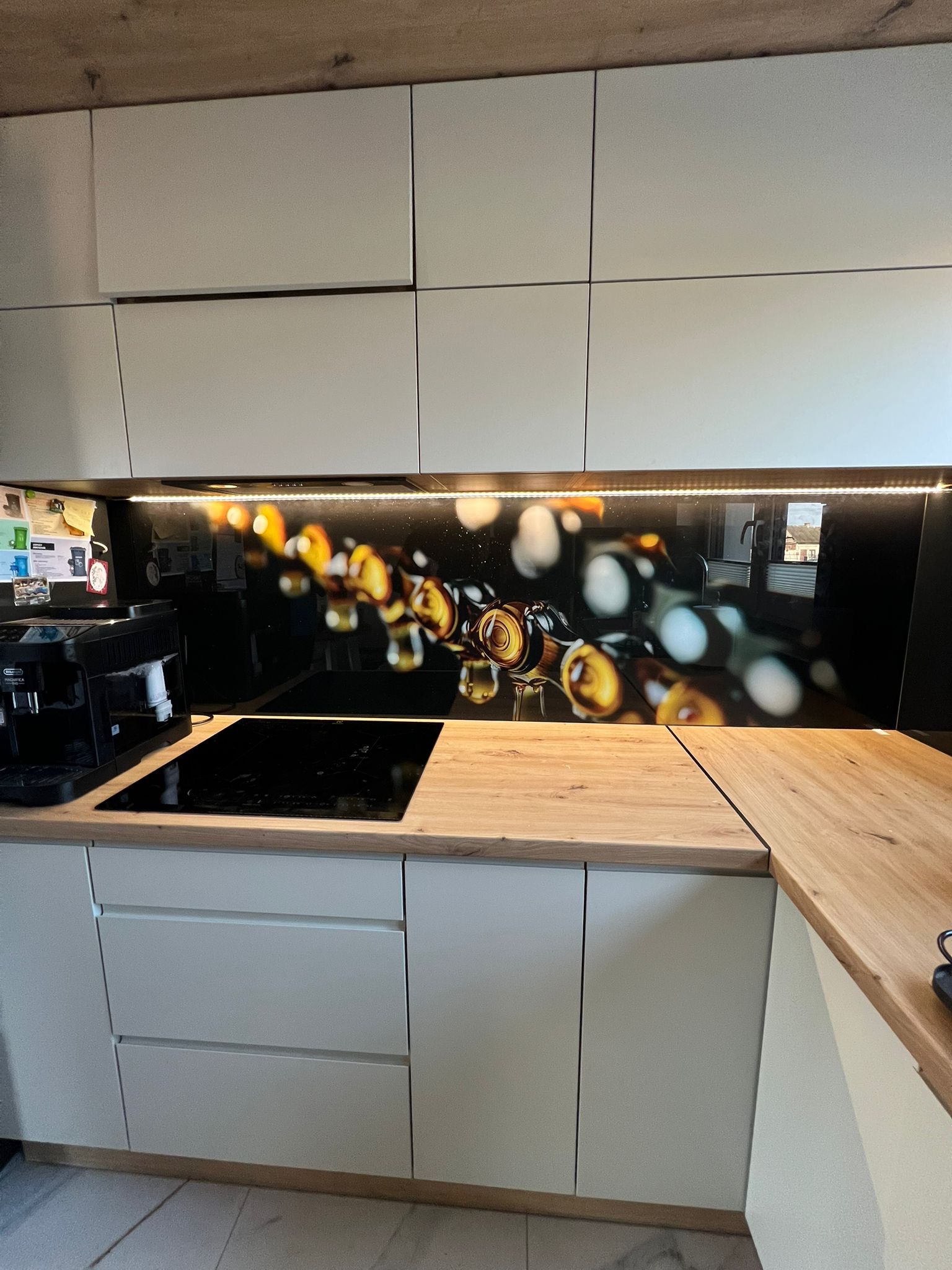 Szklany panel dekoracyjny z motywem abstrakcyjnych butelek w kuchni z białymi szafkami i drewnianym blatem, oświetlony od góry listwą LED.