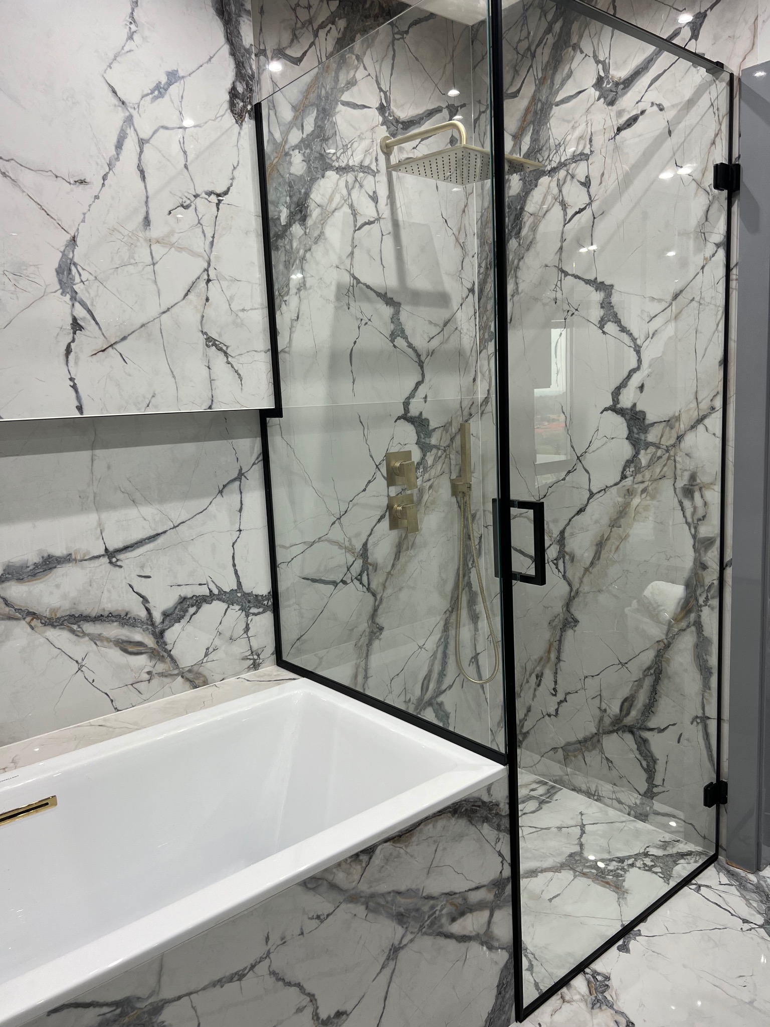Nowoczesna kabina prysznicowa ze szklanymi drzwiami w czarnej ramie, zintegrowana z białą wanną na tle marmurowych płytek
