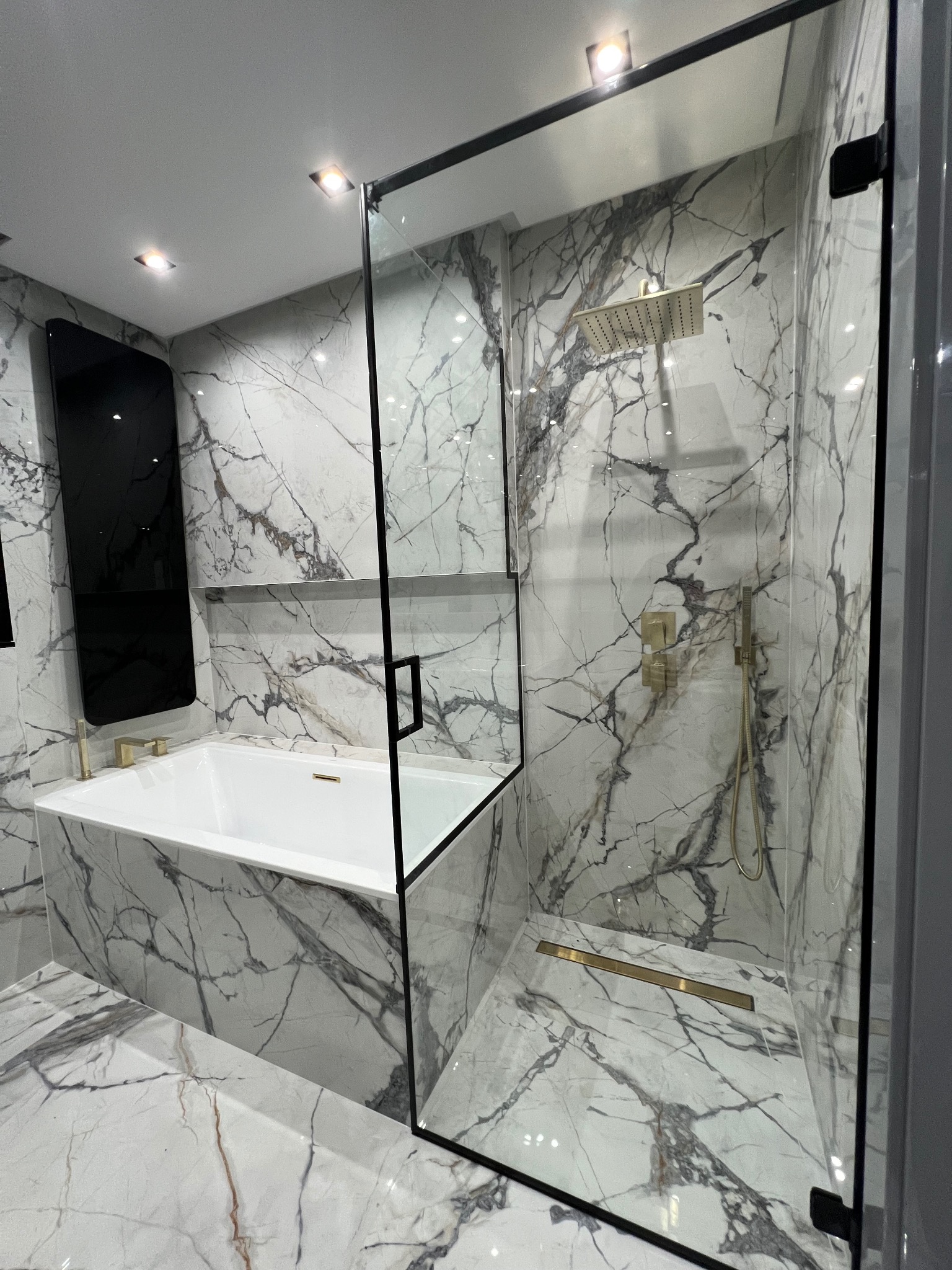 Nowoczesna łazienka z kabiną prysznicową ze szkła hartowanego z czarnymi ramami, marmurowymi ścianami i złotą armaturą, widoczna wanna obudowana marmurem.