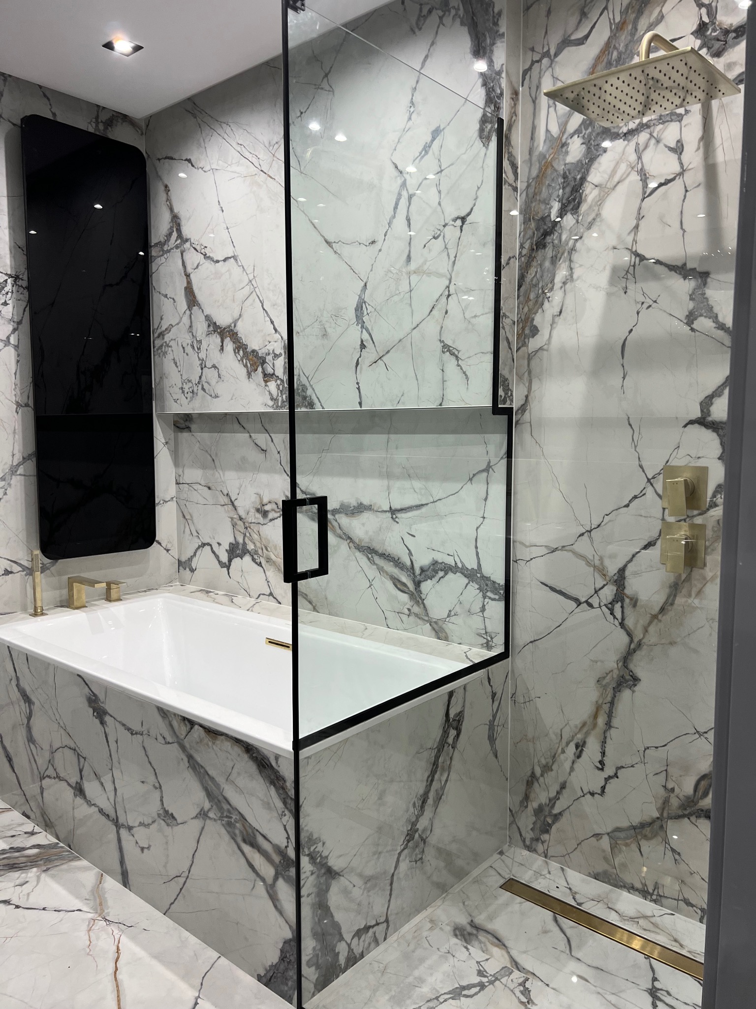 Nowoczesna łazienka z wanną, złotą armaturą i kabiną prysznicową ze szklaną ścianką, wykończona marmurem z wyraźnym wzorem.