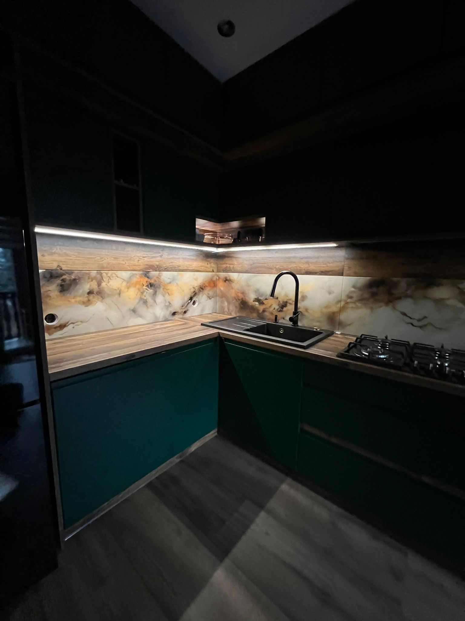 Nowoczesna kuchnia z ciemnozielonymi szafkami i blatem imitującym drewno, z podświetleniem LED nad blatem i ozdobną ścianą nad blatem w odcieniach brązu i beżu.