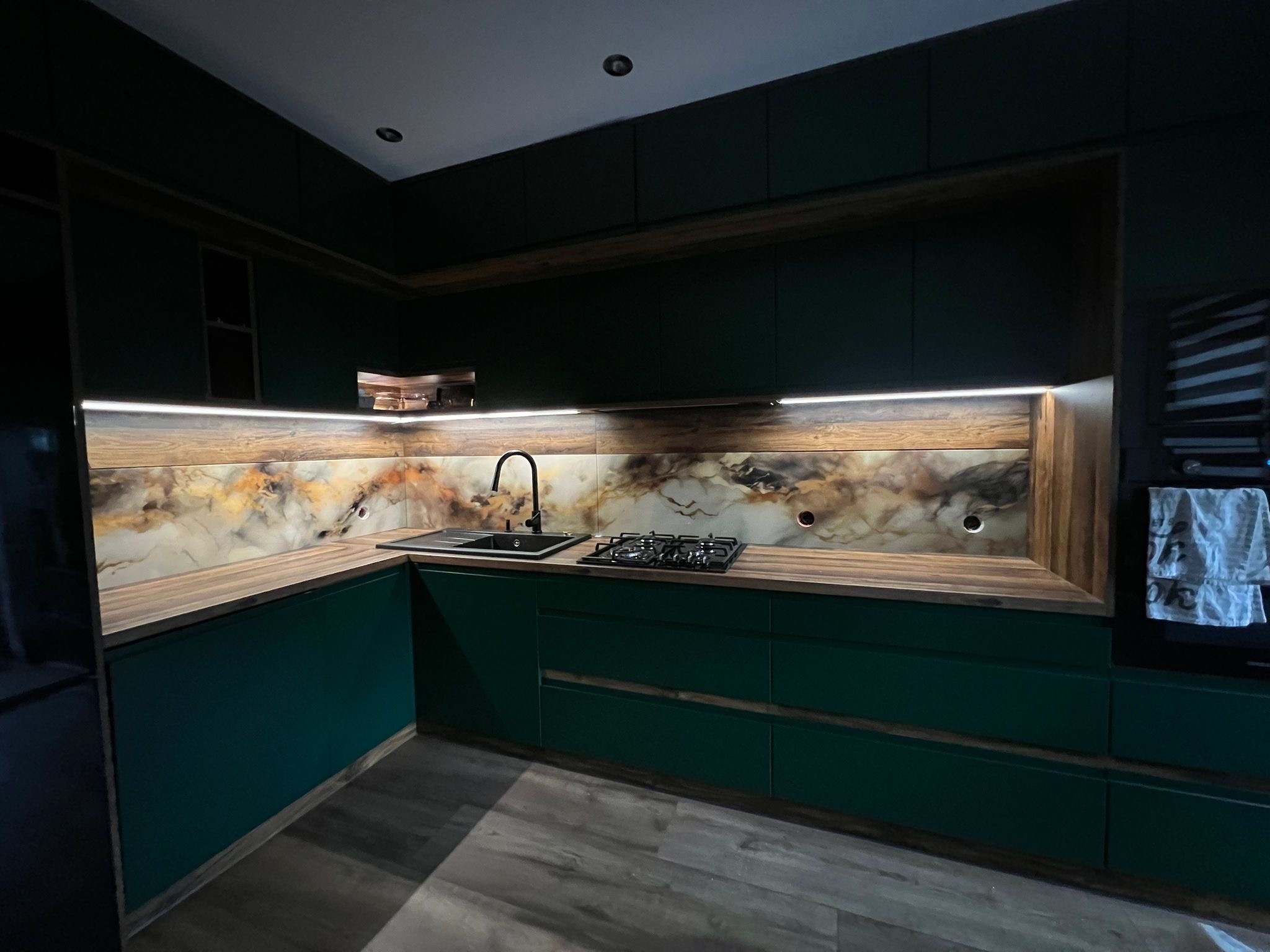 Nowoczesna kuchnia z ciemnozielonymi szafkami, drewnianym blatem i marmurkowym panelem nad blatem, oświetlona listwą LED.