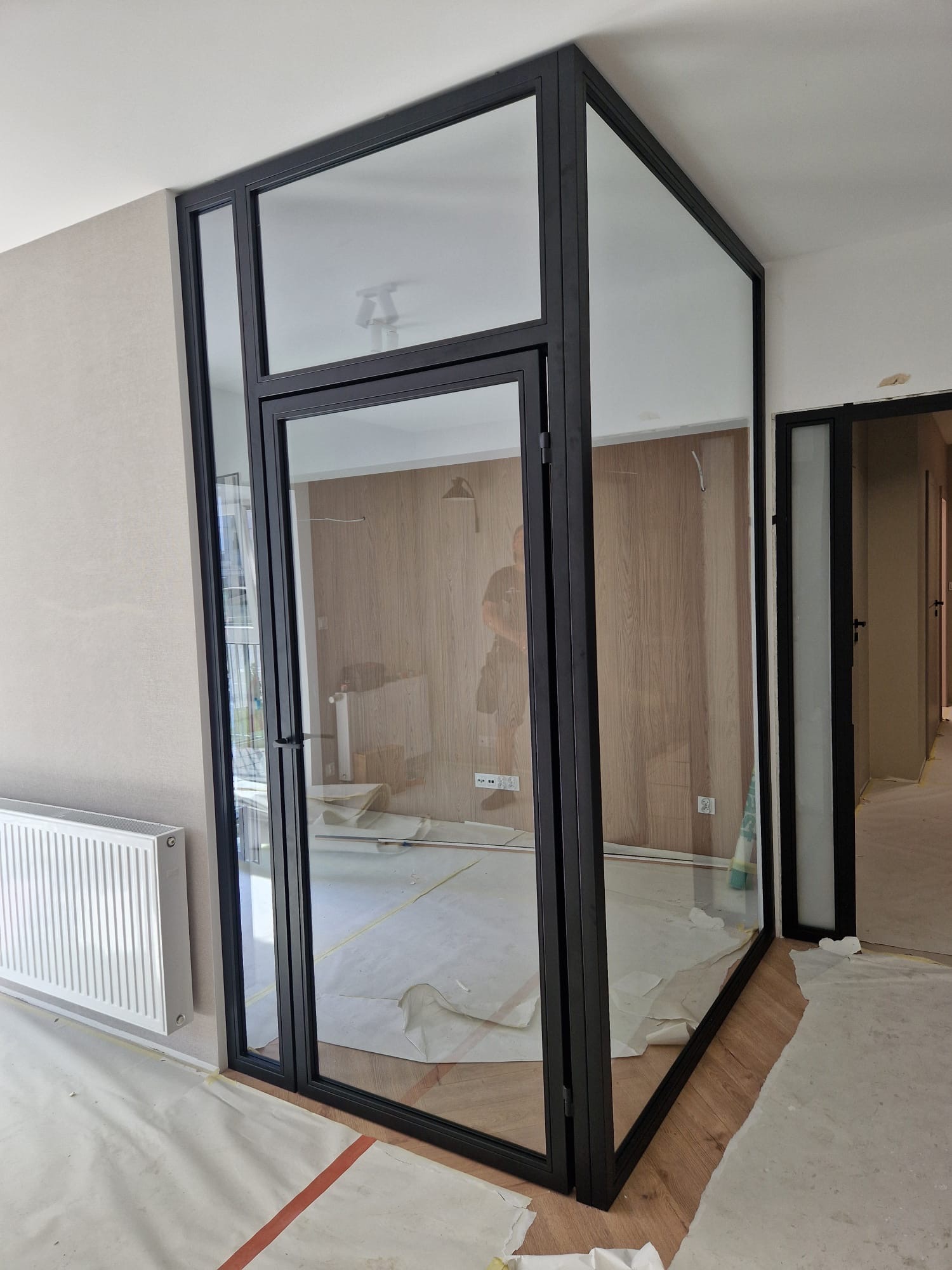 Szklana konstrukcja z czarnymi profilami tworząca ścianę działową z drzwiami, widoczna w trakcie montażu, z zabezpieczoną podłogą i grzejnikiem w tle.