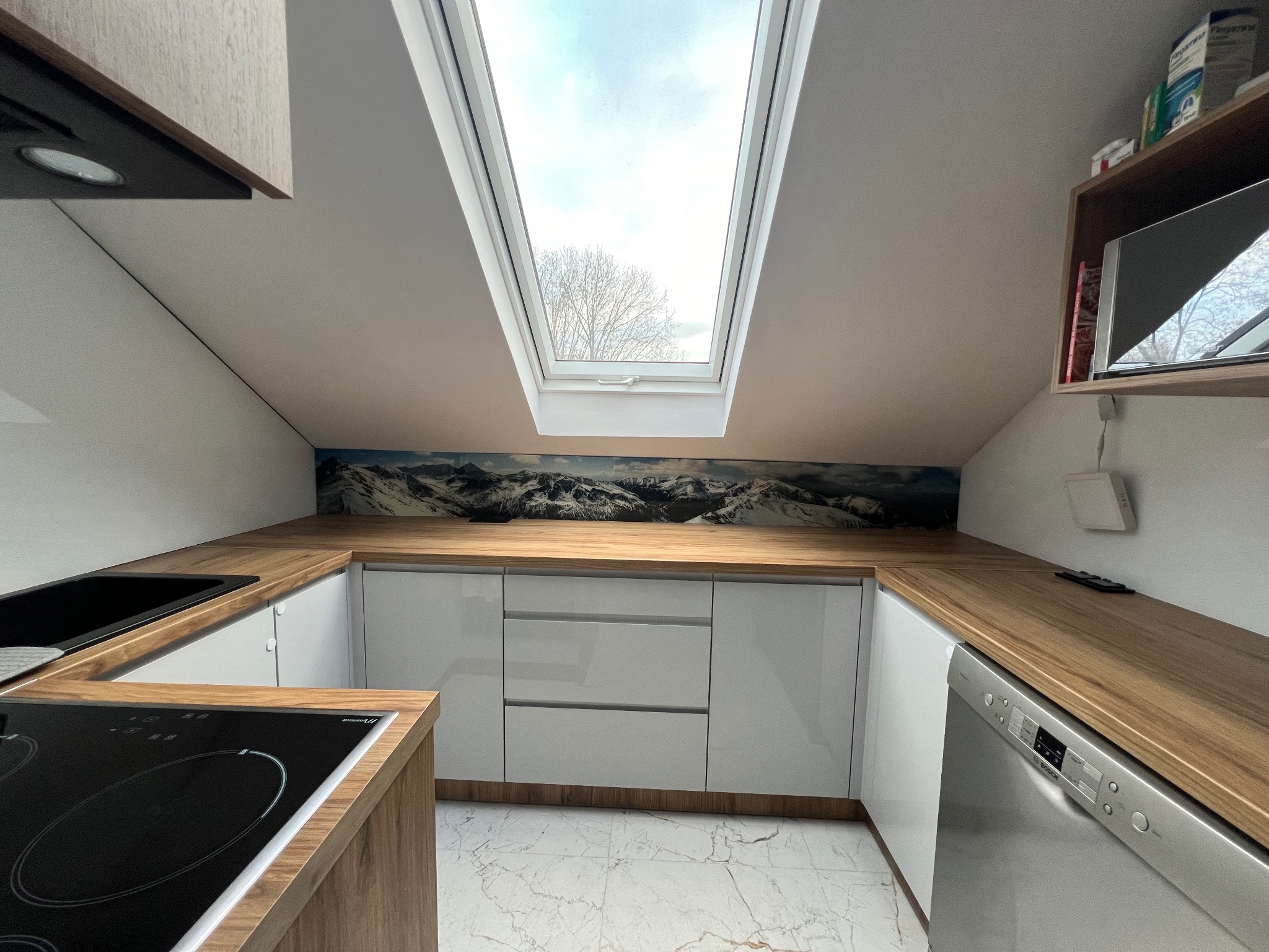 Nowoczesna kuchnia na poddaszu z białymi szafkami, drewnianym blatem i oknem dachowym z widokiem na drzewo.