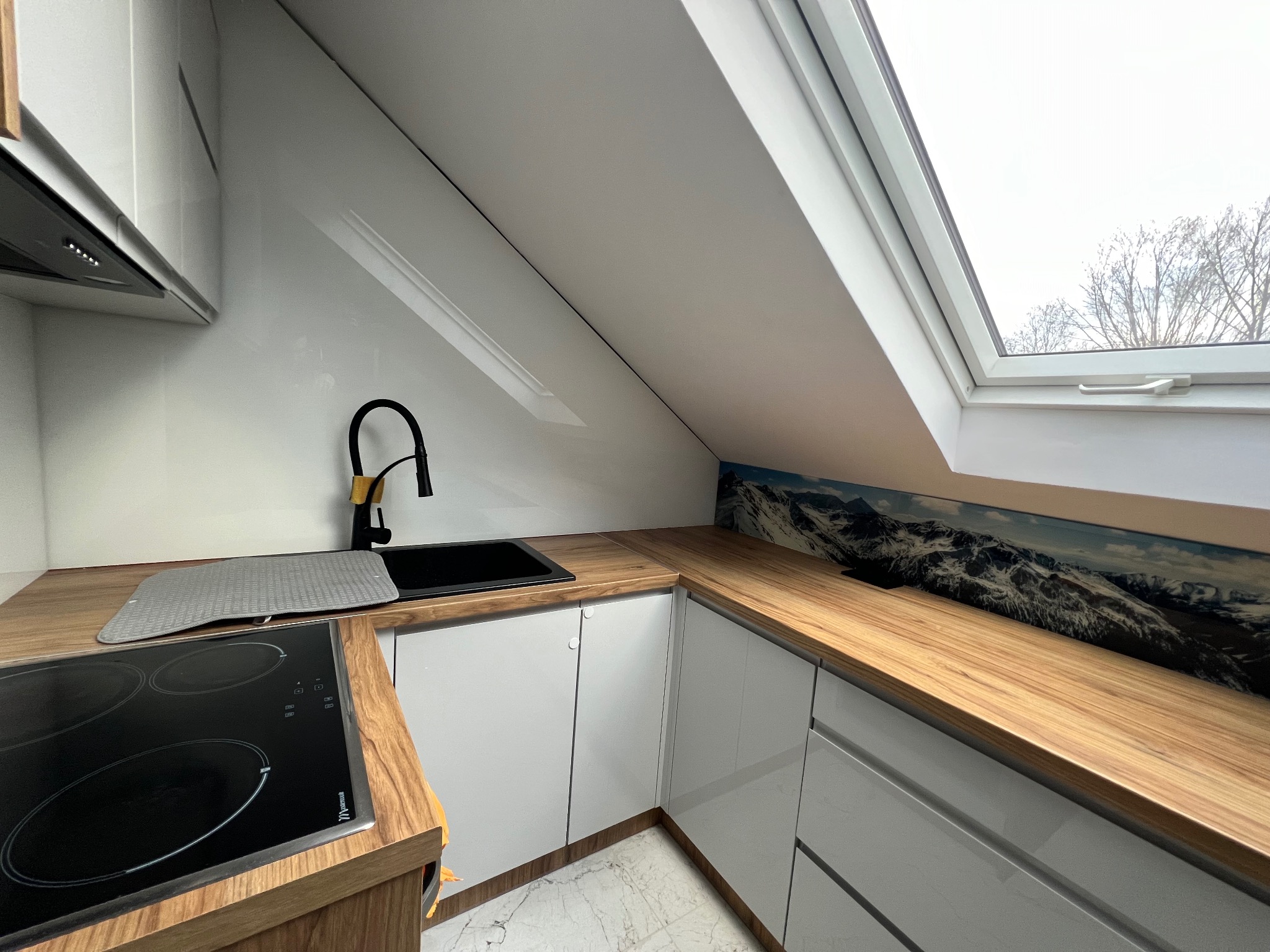 Nowoczesna kuchnia na poddaszu z białymi szafkami, drewnianym blatem, czarnym zlewem i płytą indukcyjną; nad blatem panel ze zdjęciem gór, a nad zlewem okno dachowe.