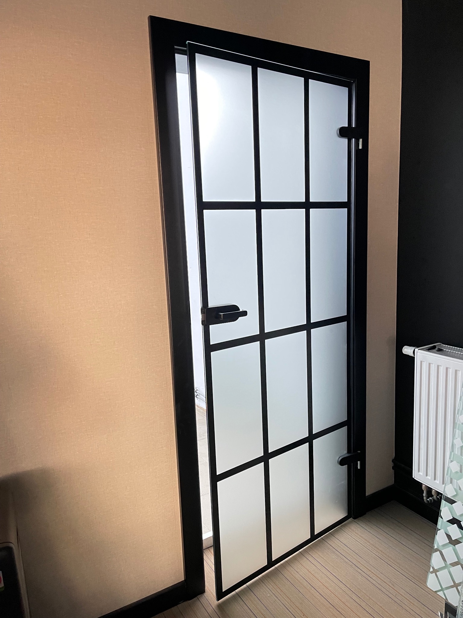 Przesuwne drzwi z matowego szkła w czarnej ramie, podzielone na prostokątne pola, widoczne w otworze ściany o dwóch kolorach.