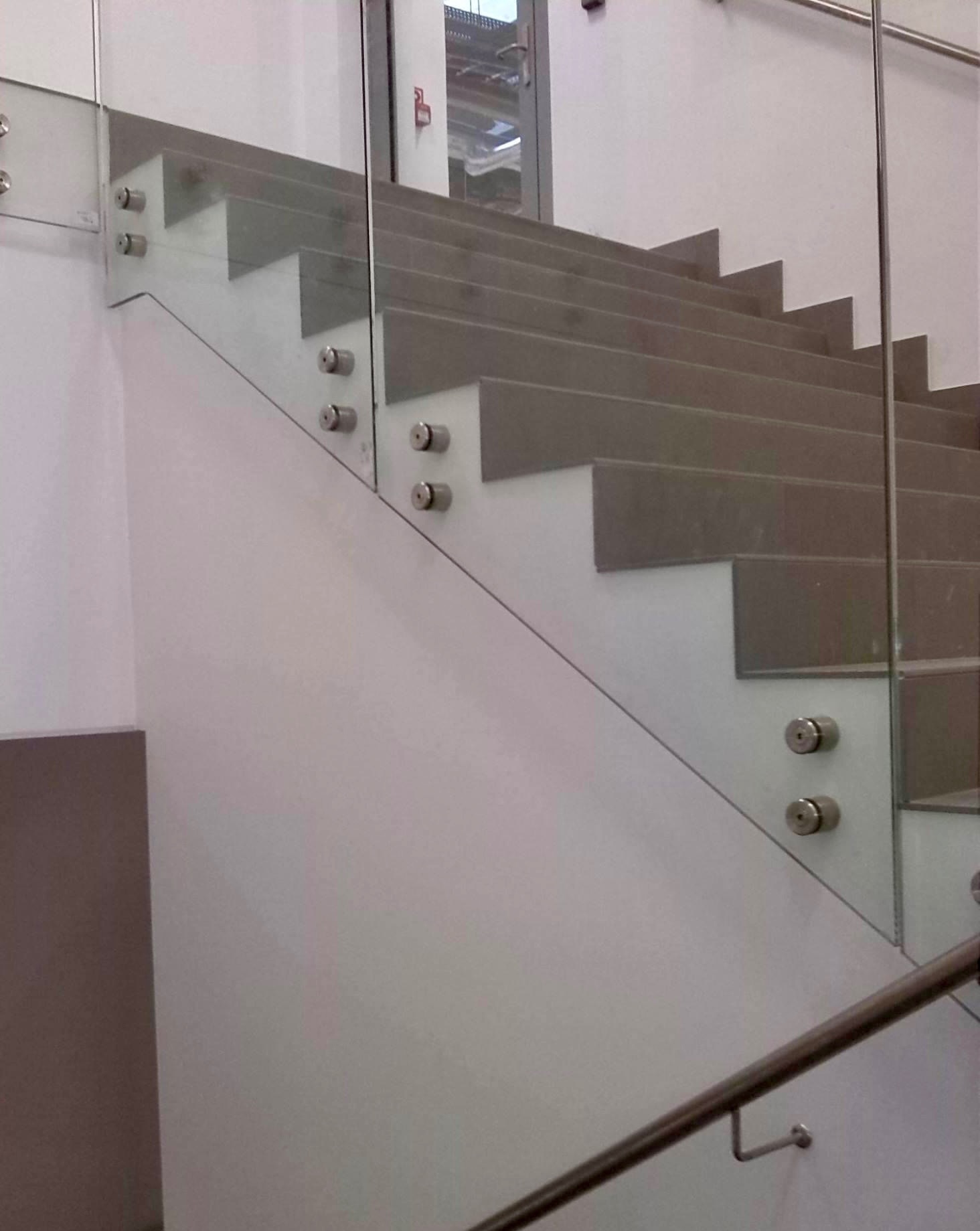 Schody z szarego kamienia zabezpieczone szklaną balustradą mocowaną punktowo, widoczne metalowe poręcze.