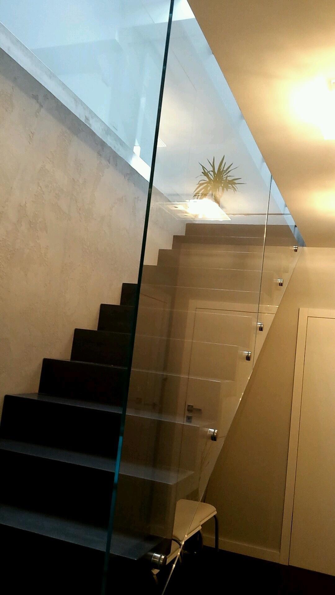 Nowoczesne schody z czarnymi stopniami zabezpieczone szklaną balustradą mocowaną punktowo, widok na górny poziom z rośliną doniczkową.