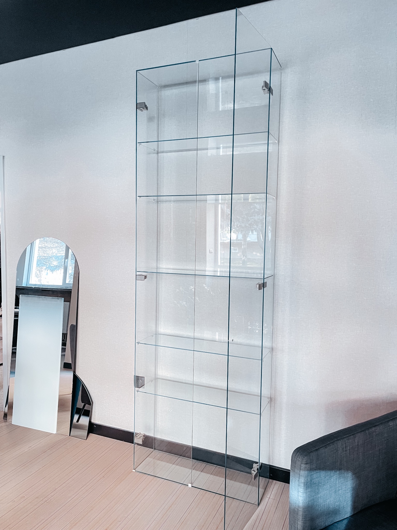 Przezroczysta, szklana witryna z półkami i drzwiczkami, zamontowana w nowoczesnym wnętrzu, obok lustra w kształcie łuku.