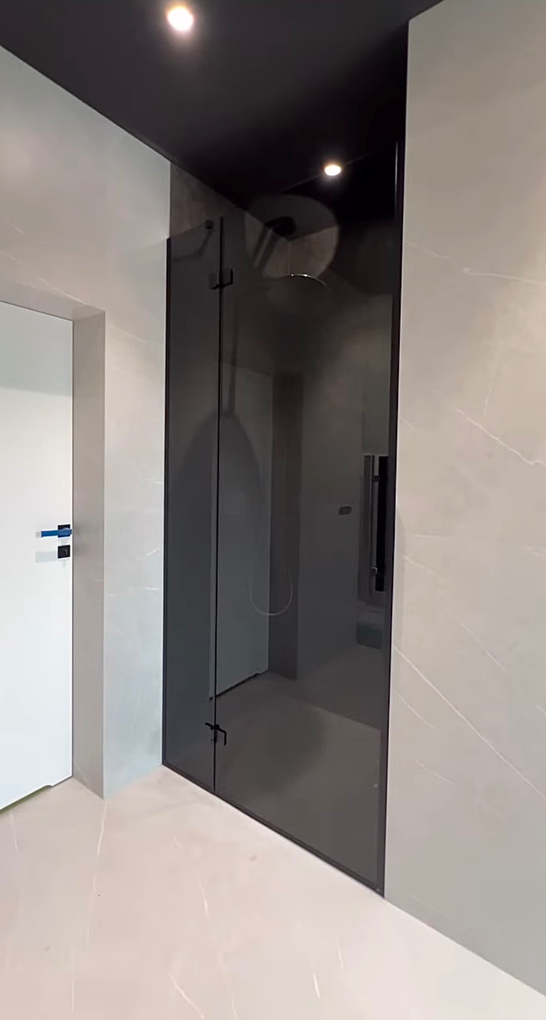 Nowoczesna kabina prysznicowa z ciemnego szkła, widoczna armatura i deszczownica, białe marmurowe płytki na ścianach i podłodze, oświetlenie punktowe.